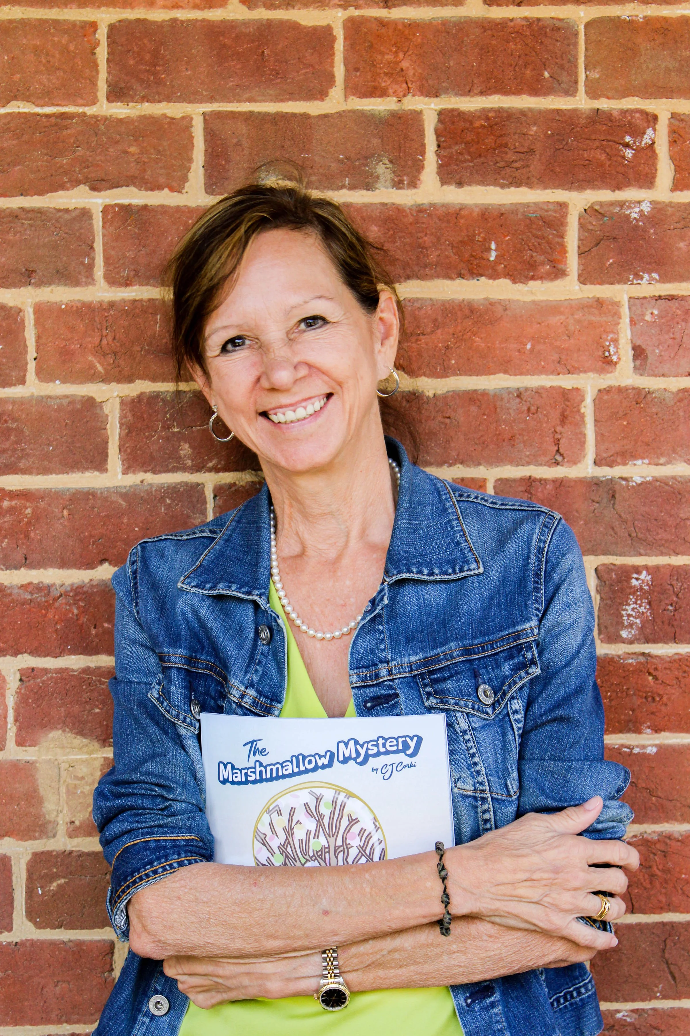 A smiling woman with short brown hair, wearing hoop earrings, a pearl necklace, a denim jacket, and a yellow top, standing against a brick wall while holding a book titled 'The Marshmallow Mystery' by J.C. Cook.