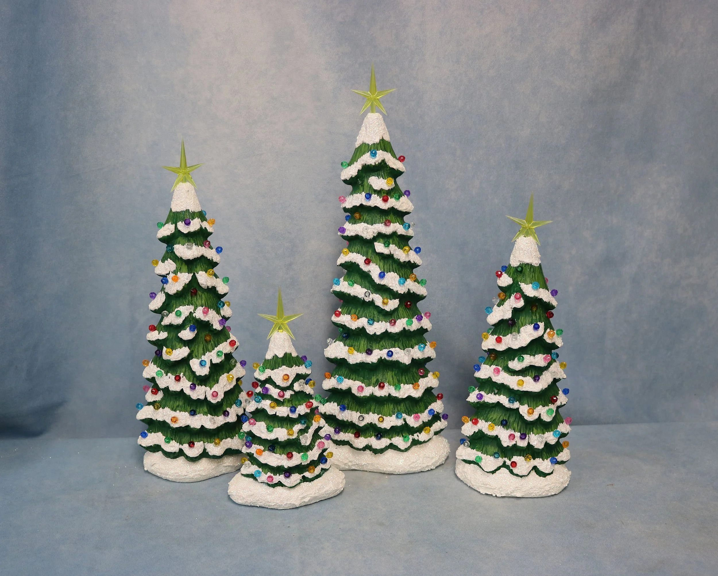 Four small Christmas trees decorated with multicolored ornaments and white garlands, topped with yellow stars, set against a blue background.