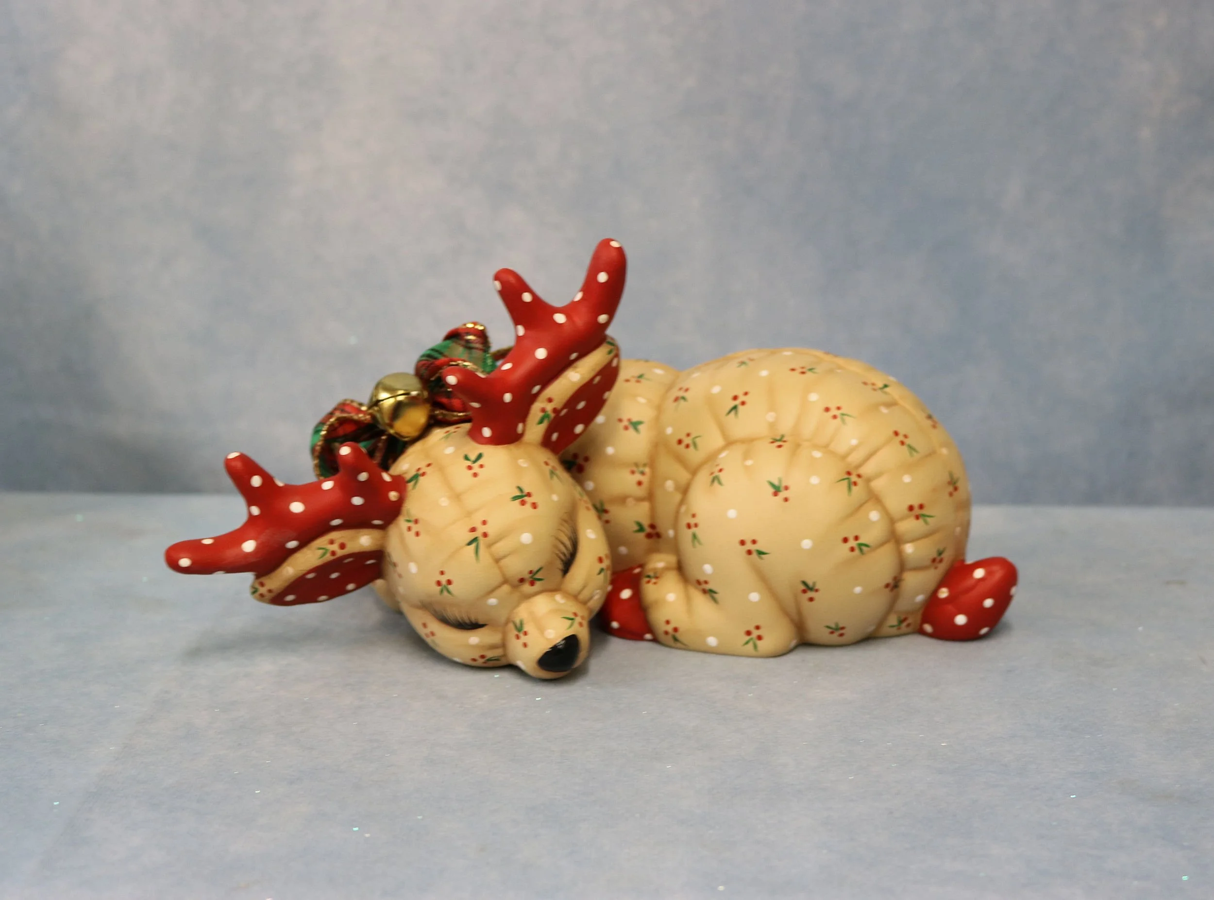 Decorative ceramic deer with antlers, painted in red with white polka dots, resting with small red and green floral patterns, set against a plain blue background.