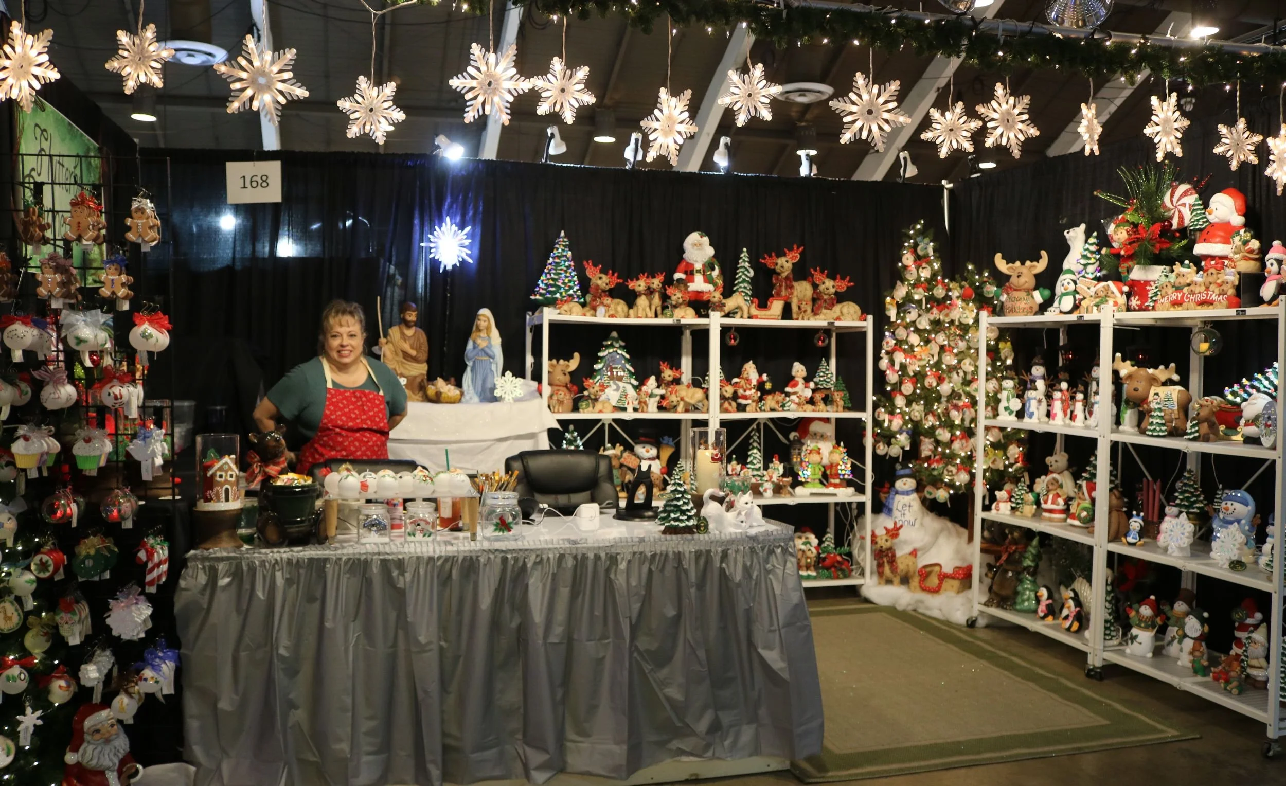 Christmas market booth decorated with holiday lights, snowflake ornaments hanging from the ceiling, and shelves displaying Christmas-themed decorations like Santa figurines, reindeer, snowmen, and ornaments. A vendor woman stands behind the table smiling.