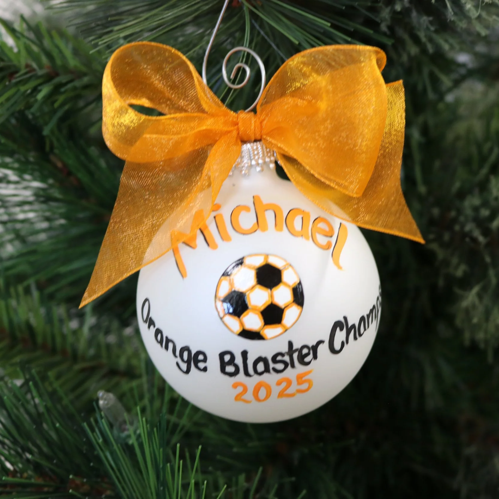 A white ornament on a Christmas tree decorated with a large gold ribbon bow, with the words 'Orange Blaster Champ 2025' and a soccer ball graphic.