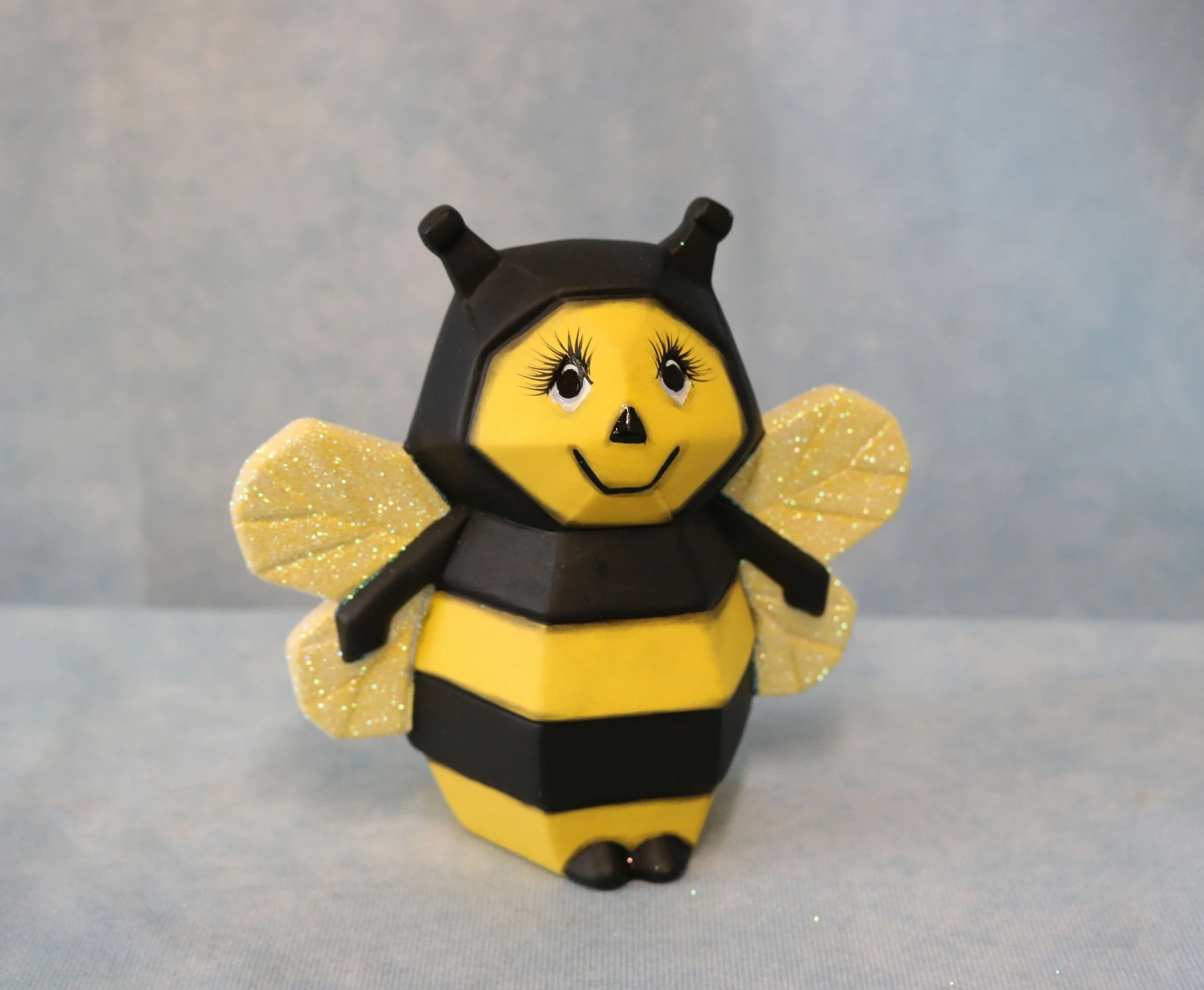 A small decorative bee with a yellow face, large eyes with long eyelashes, a black nose, and a smiling mouth. It has black and yellow body stripes, glittery yellow wings, and black antennae, set against a light gray background.