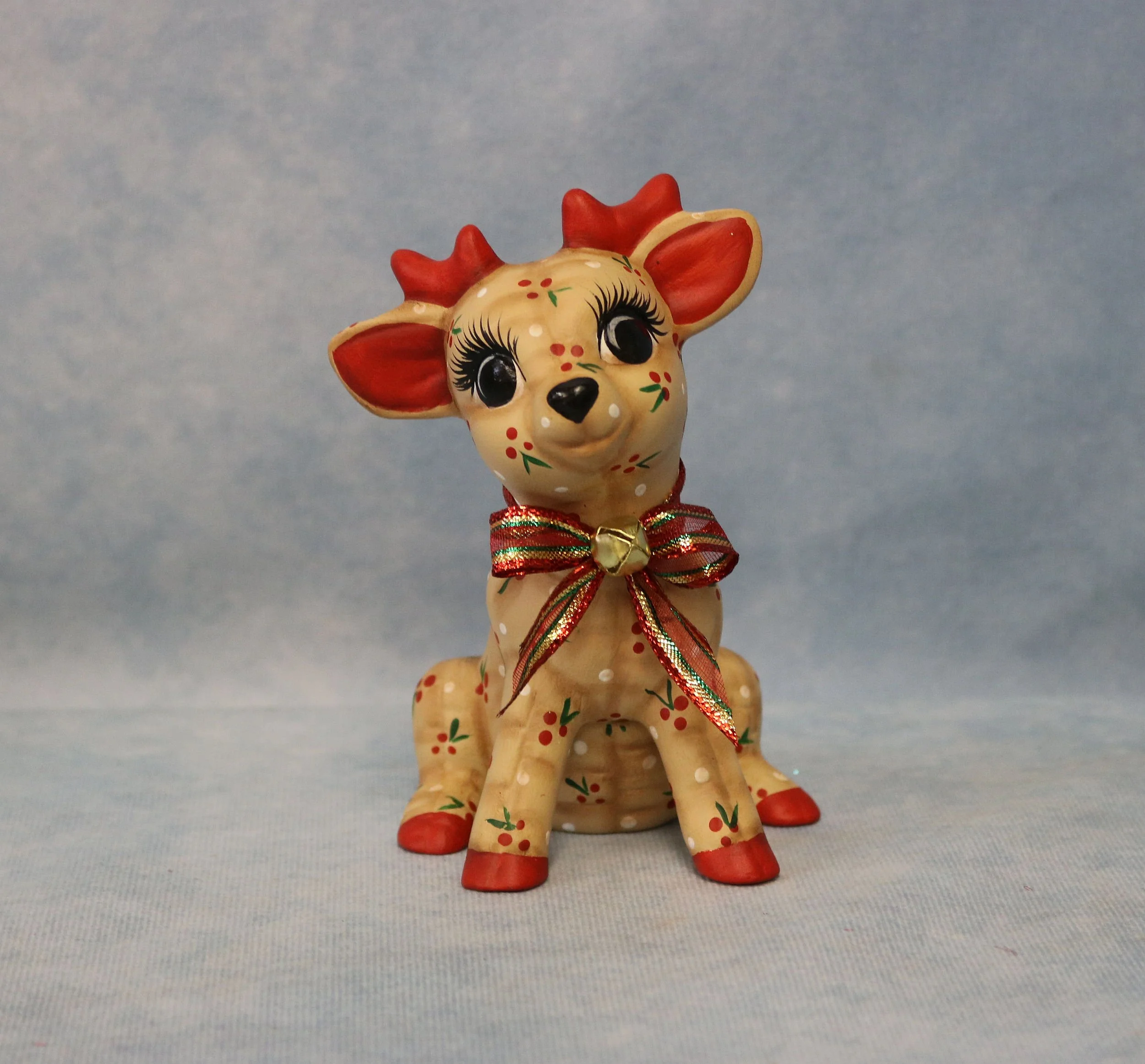 A small painted reindeer with red and green decorations, wearing a festive red and green striped ribbon around its neck, standing on a neutral surface with a plain blue background.