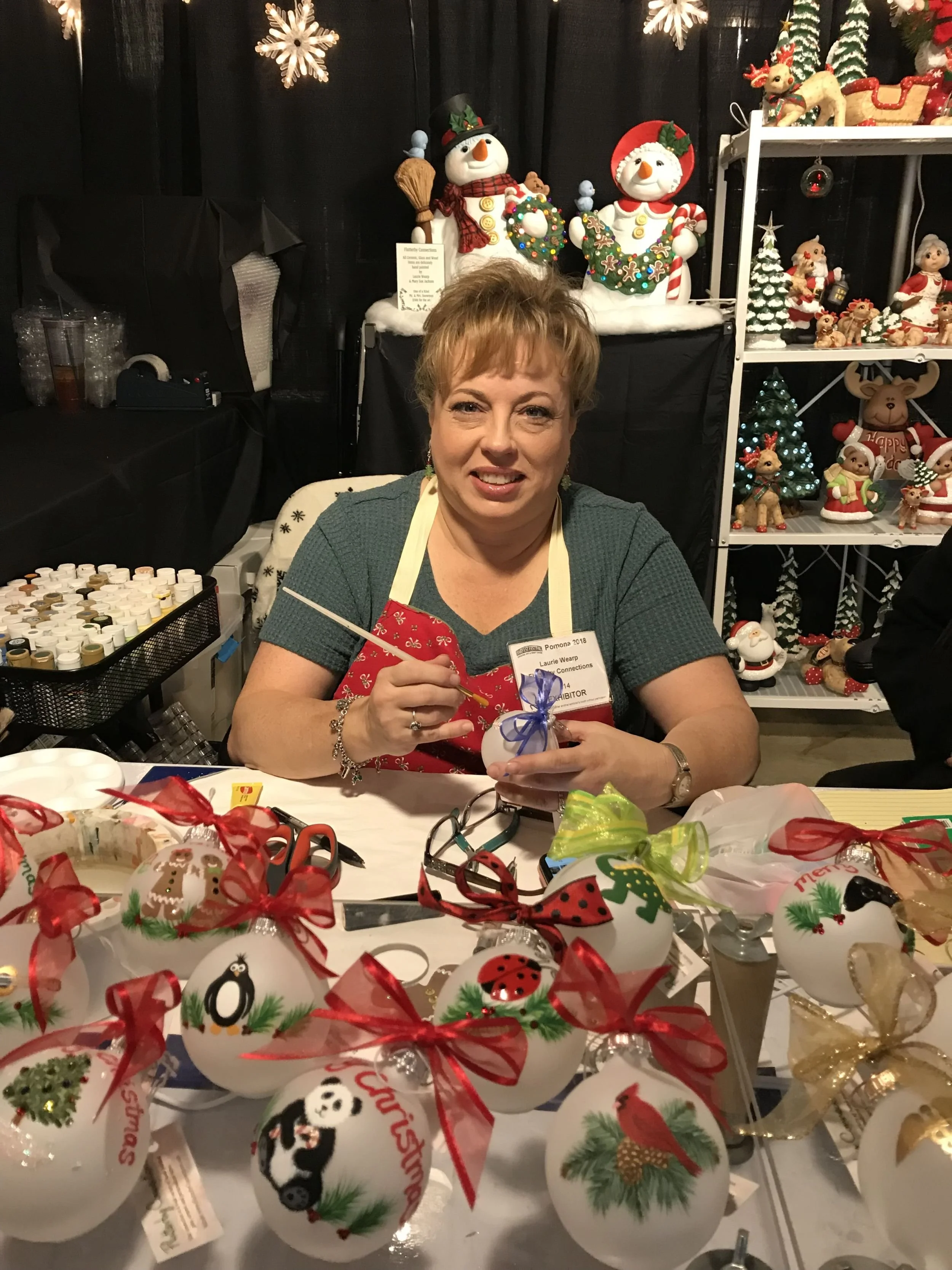 A woman sitting at a table painting personalized Christmas ornaments and decorations, with holiday themes and bows, set against a background of Christmas figurines and decorations on shelves.