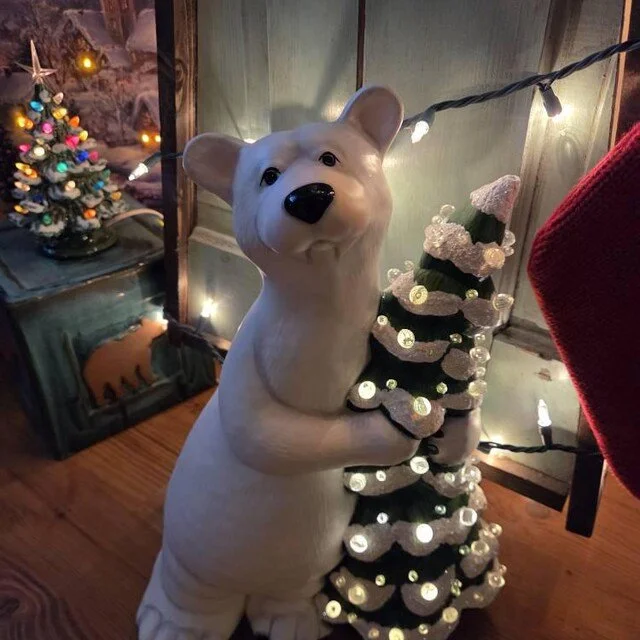 Decorative polar bear sculpture holding a small, decorated Christmas tree with string lights around it, and a small decorated Christmas tree with colorful lights in the background.