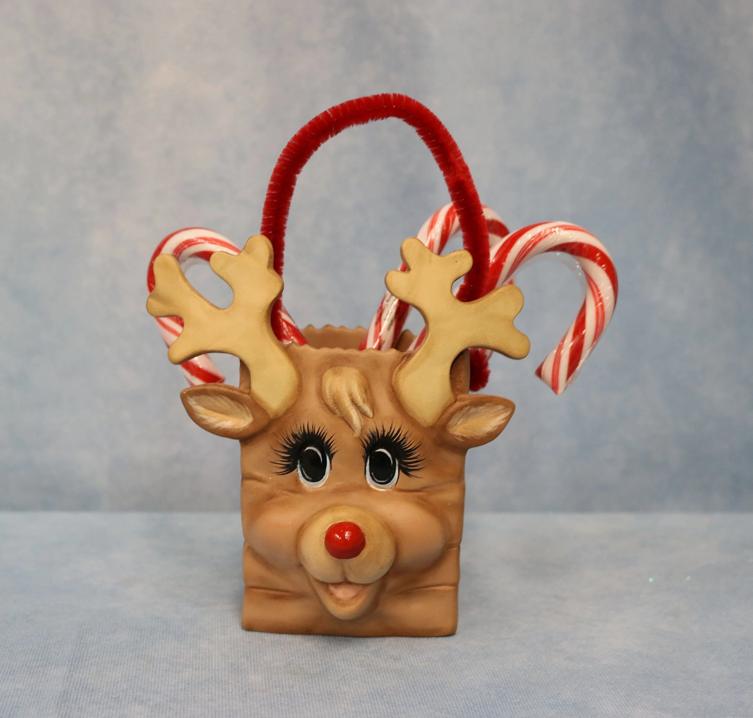 Decorative reindeer ceramic with candy canes, red nose, and large eyes, set against a light blue background.