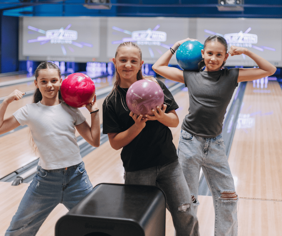 Three girls standing by Play2Win Bowling lanes holding bowling balls in their hands and on their shoulders