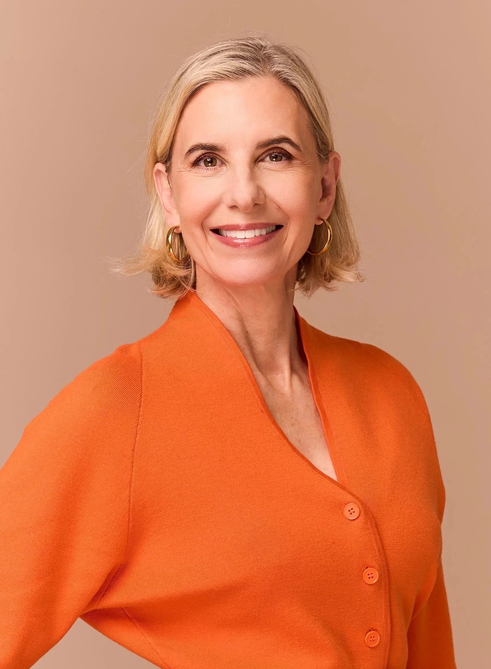 Maria Eitel. A smiling  woman with short blonde hair, wearing gold hoop earrings and an orange button-up top, standing against a neutral background.