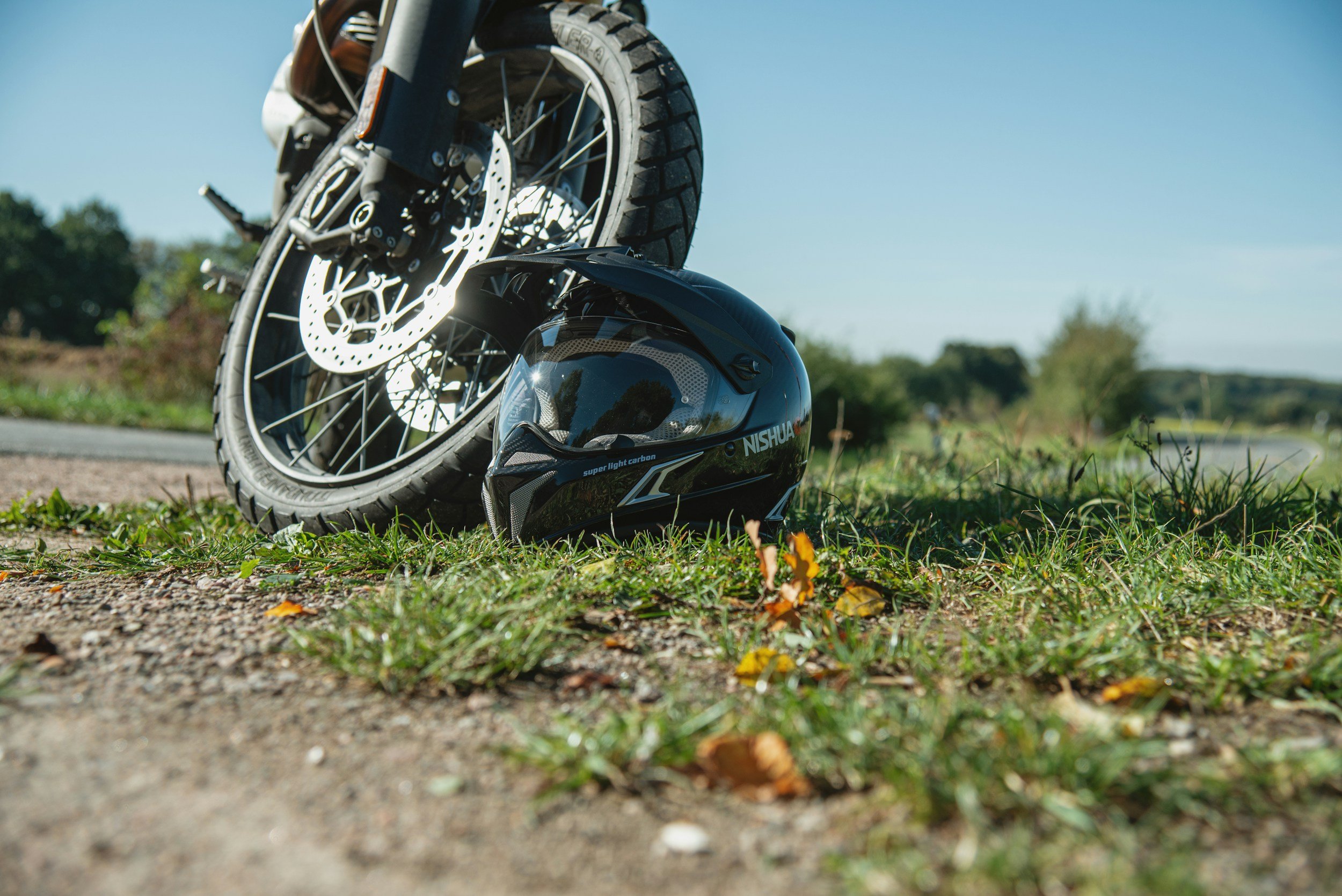 A motorcycle with a helmet resting on the ground next to it, taken outdoors on a sunny day with trees and open sky in the background.