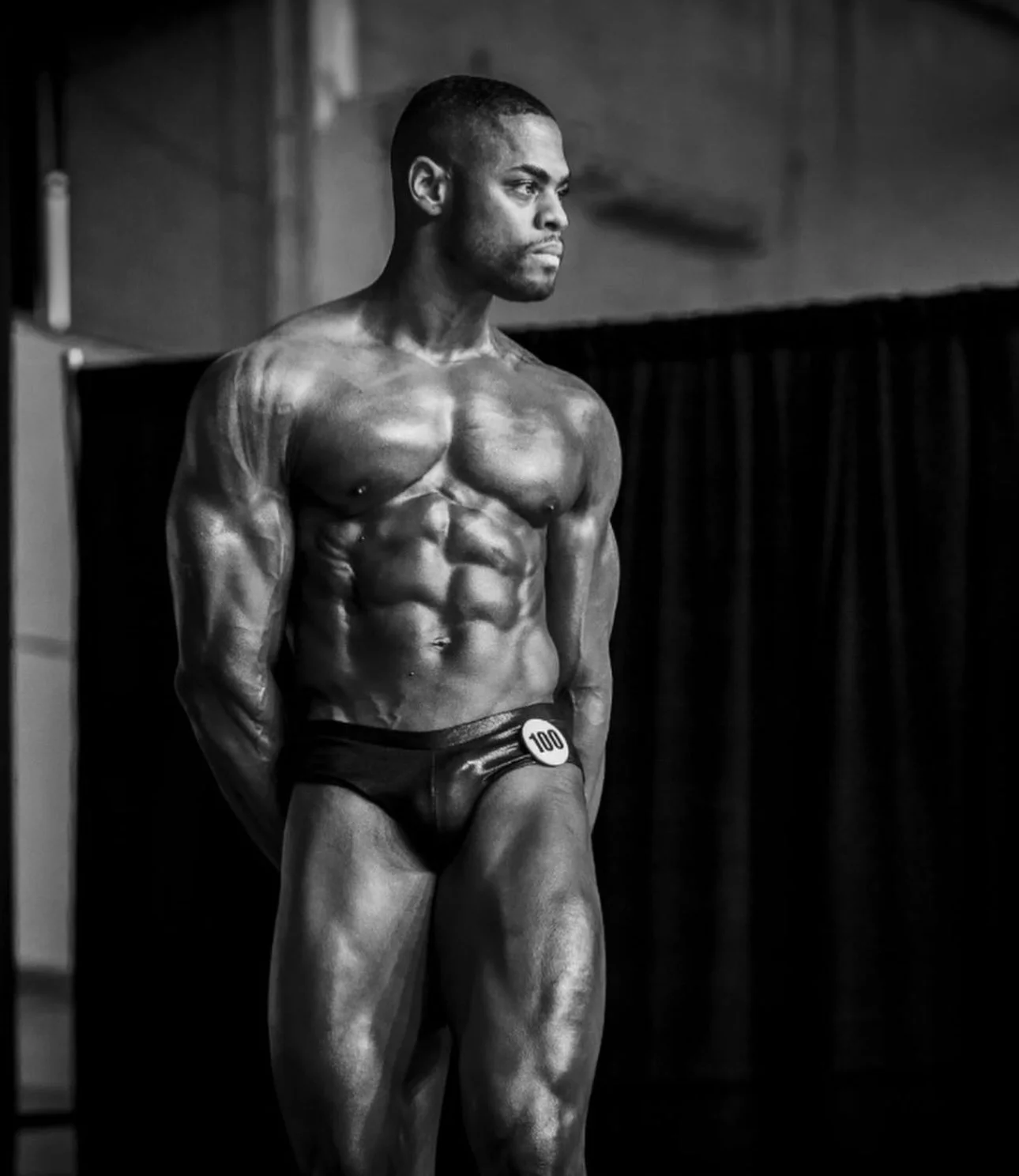 Black and white photo of a male bodybuilder on stage, wearing a competition number 100, posing to display muscular physique.