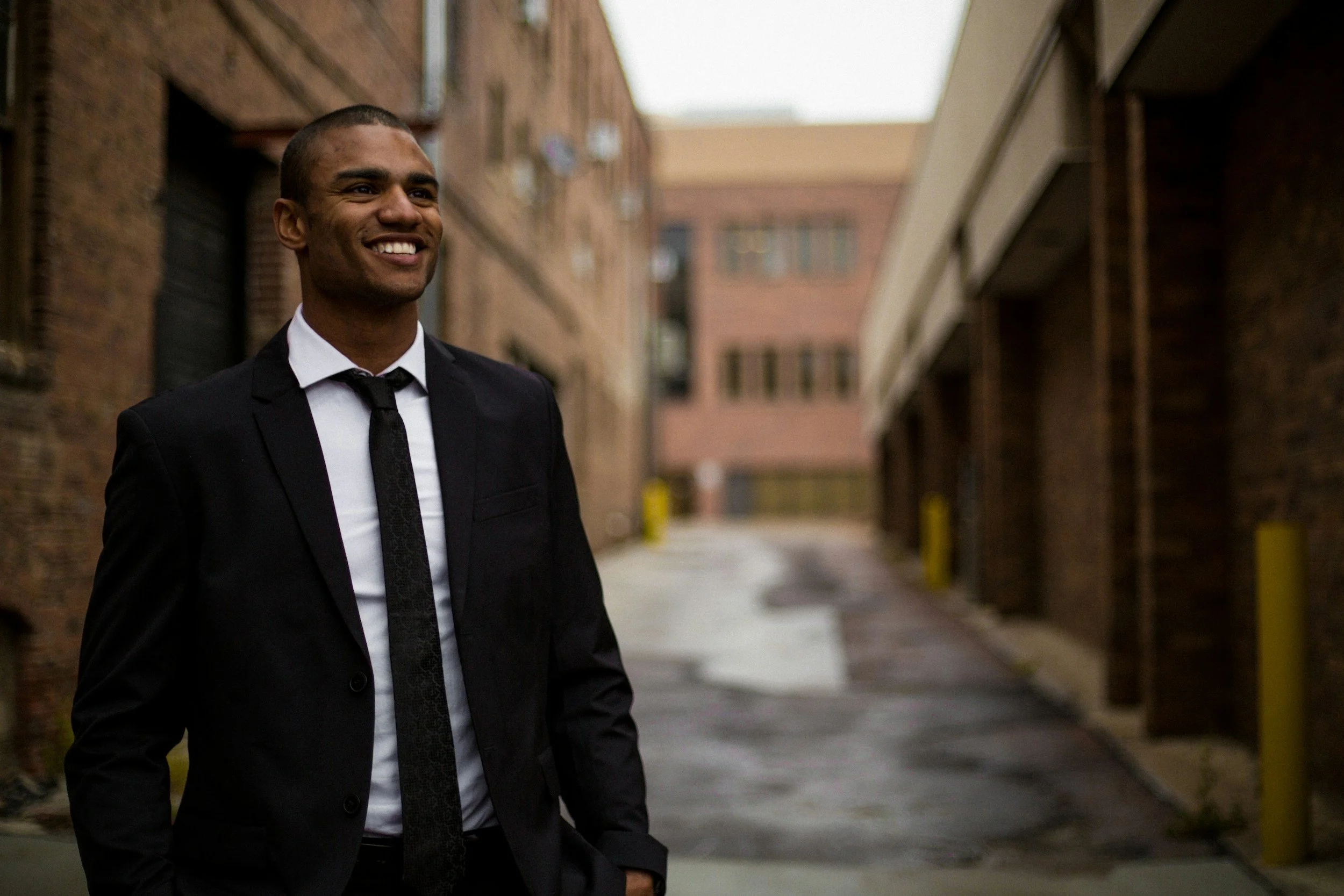 Smiling man in business suit standing in alleyway with brick building walls on either side.