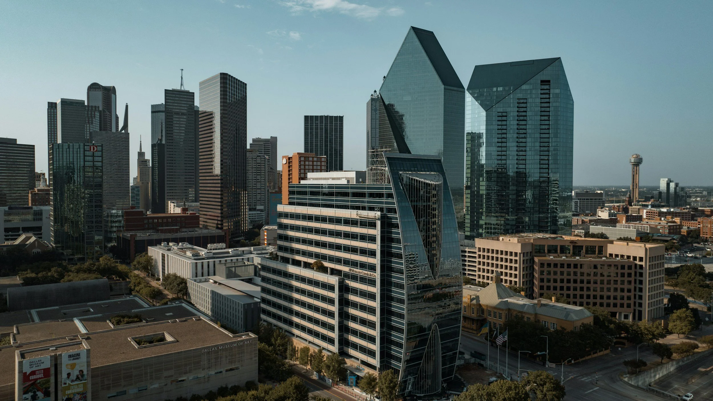A city skyline featuring tall modern glass skyscrapers, including buildings with unique geometric shapes, a tower with a spherical top, and various mid-rise buildings, under a clear sky.