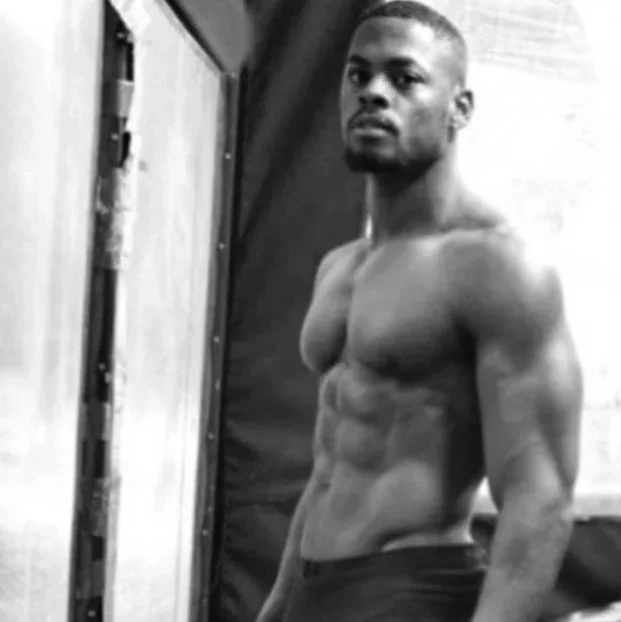 A shirtless man with a muscular physique taking a selfie in front of a mirror.