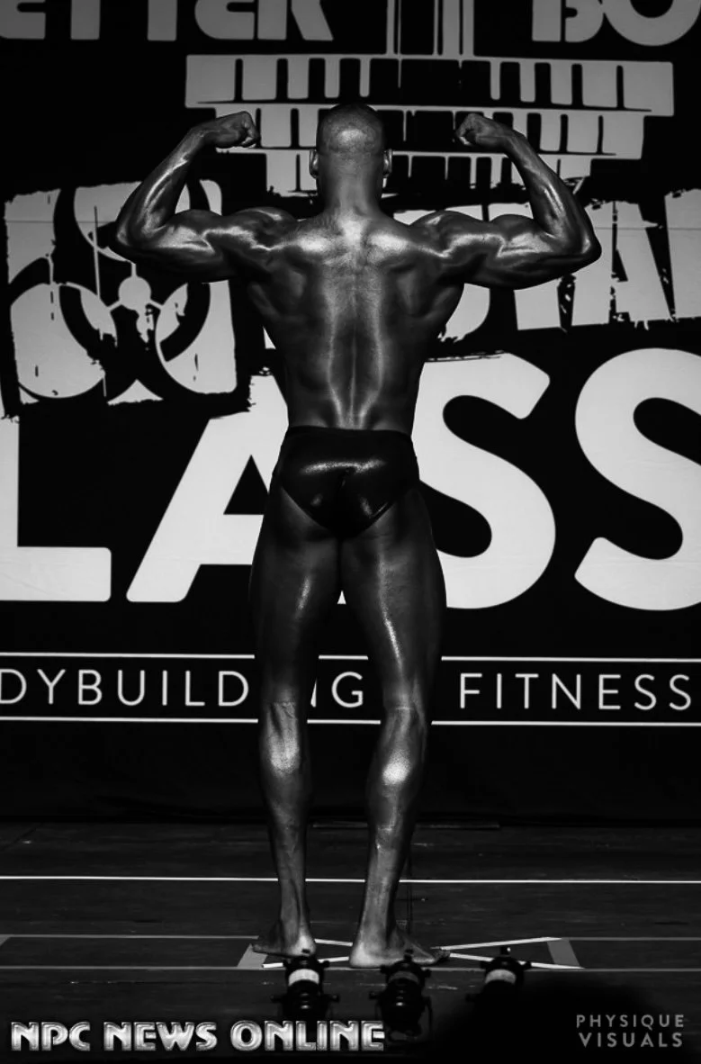 A male bodybuilder is standing on stage in a bodybuilding competition, flexing his back and arm muscles, wearing black posing trunks.