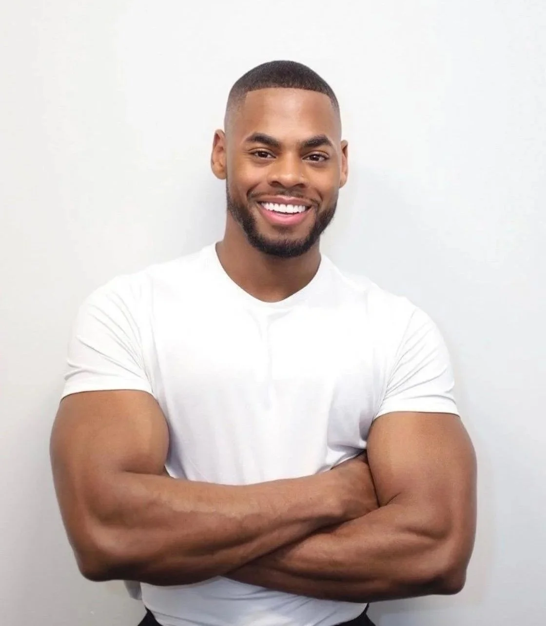 A smiling man with a beard and short hair, wearing a white t-shirt, standing with arms crossed against a plain white background.
