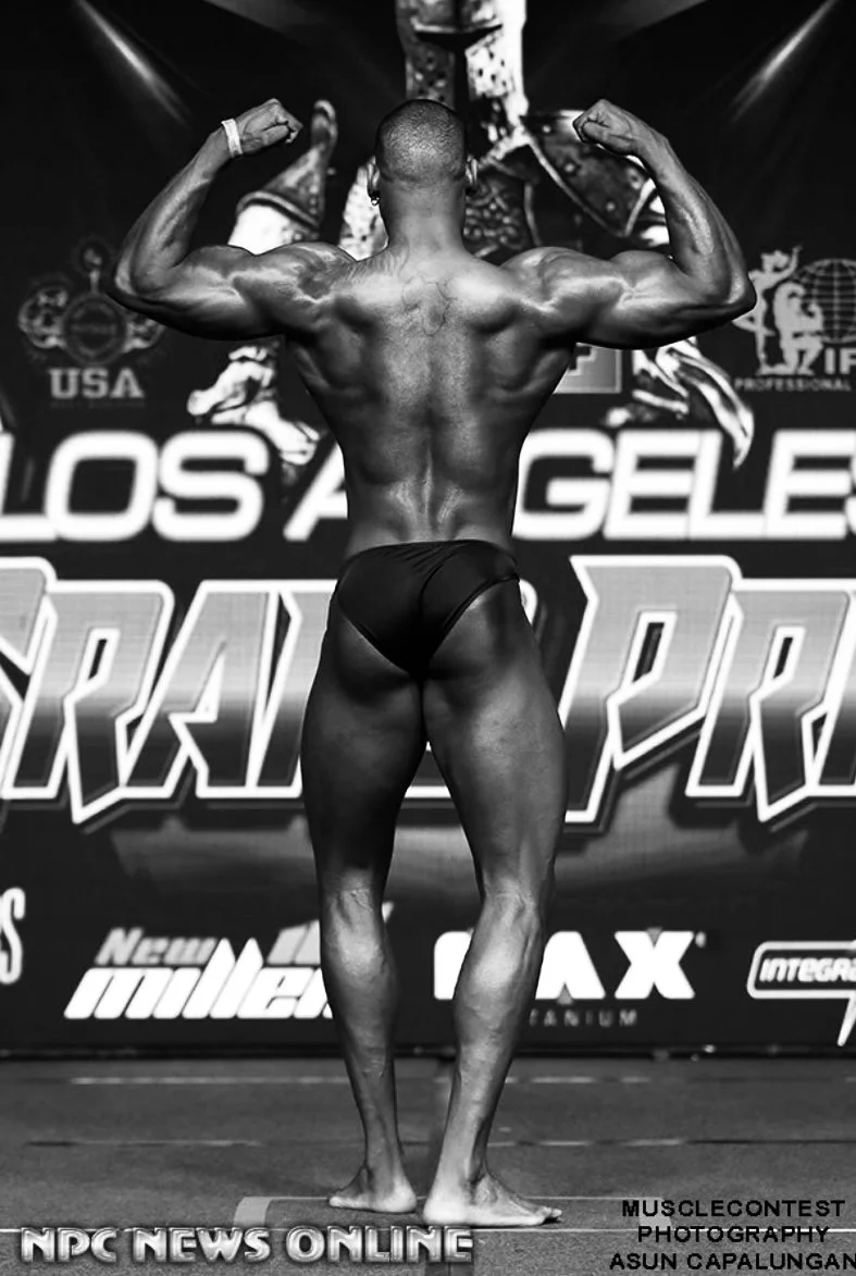 Back view of a male bodybuilder on stage flexing his muscles during a competition in front of a backdrop with logos and text.