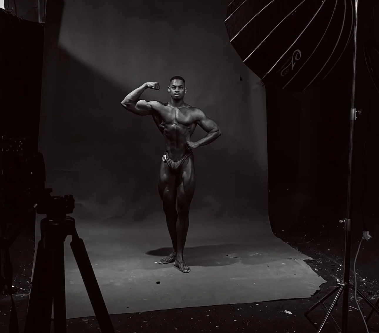 Black male bodybuilder posing in front of a gray backdrop during a photo shoot, with studio lighting and equipment visible.