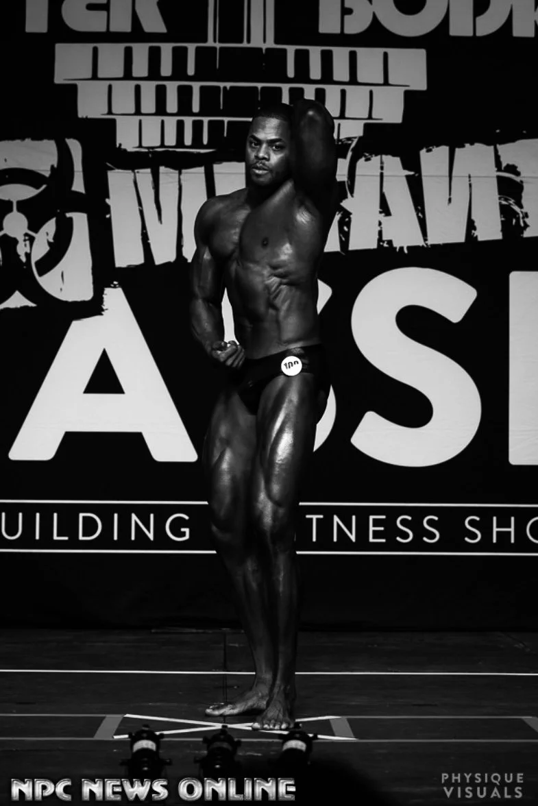 A male bodybuilder posing on stage during a competition, with a black background and large text behind him that reads "NPC News Online" and other words related to fitness. He is wearing a small posing suit and is striking a pose to showcase his muscu