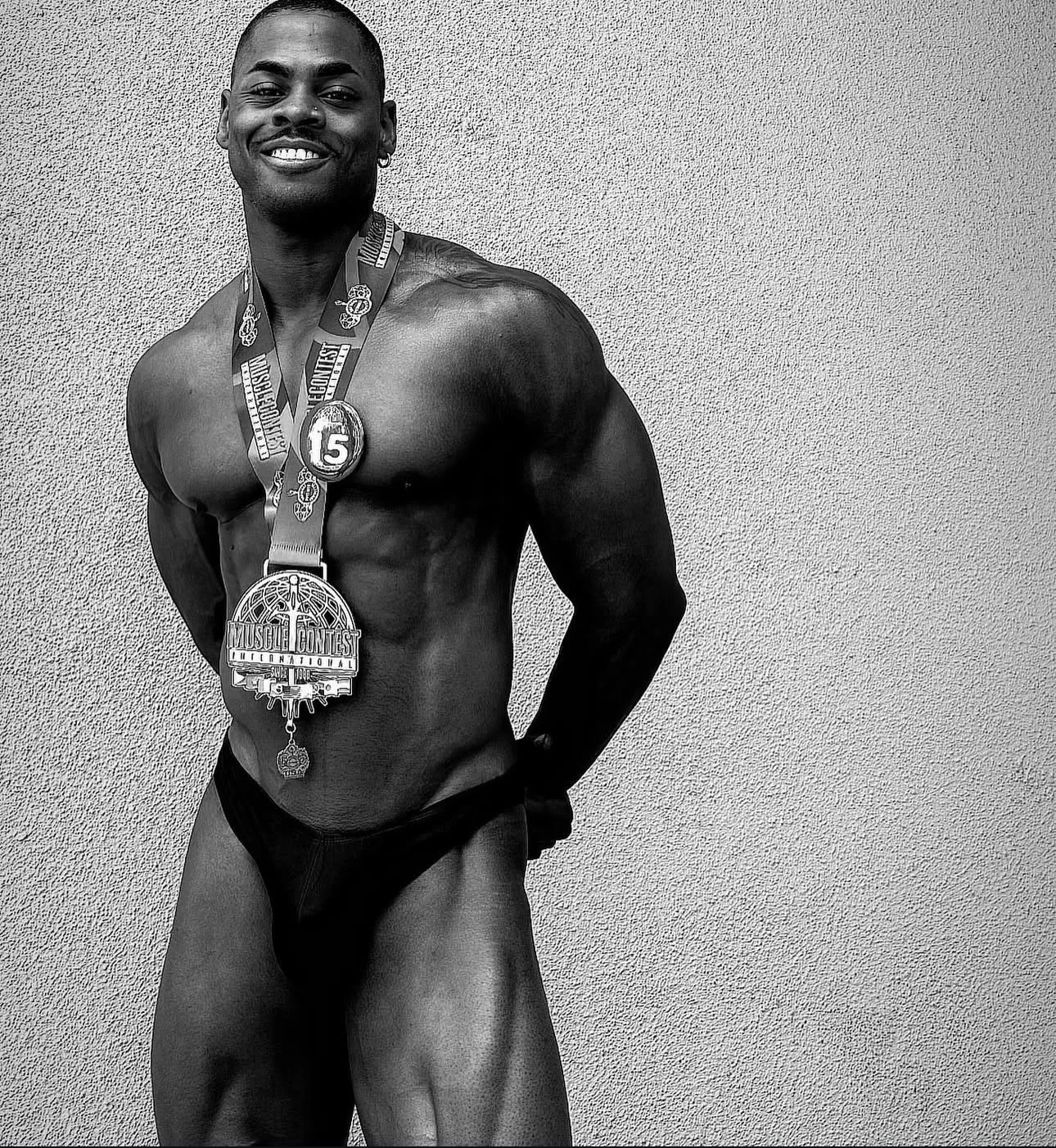 A smiling male bodybuilder with medals around his neck, wearing a small black swimsuit, standing against a plain wall.