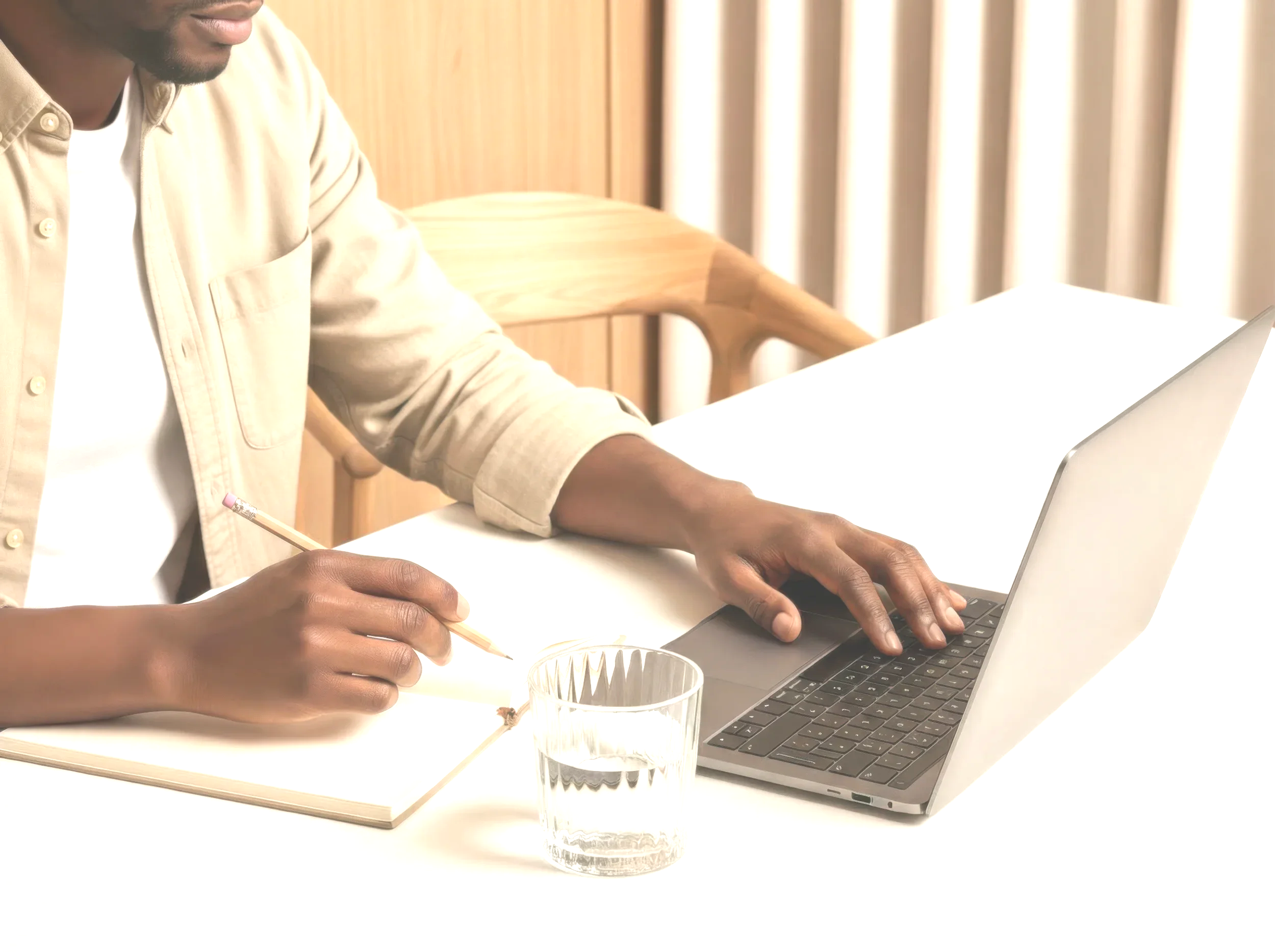 A person sitting at a white table, using a silver laptop with a black keyboard. They are holding a pencil and writing in a notebook. A glass of water is on the table, and the background features a wooden wall and curtains.