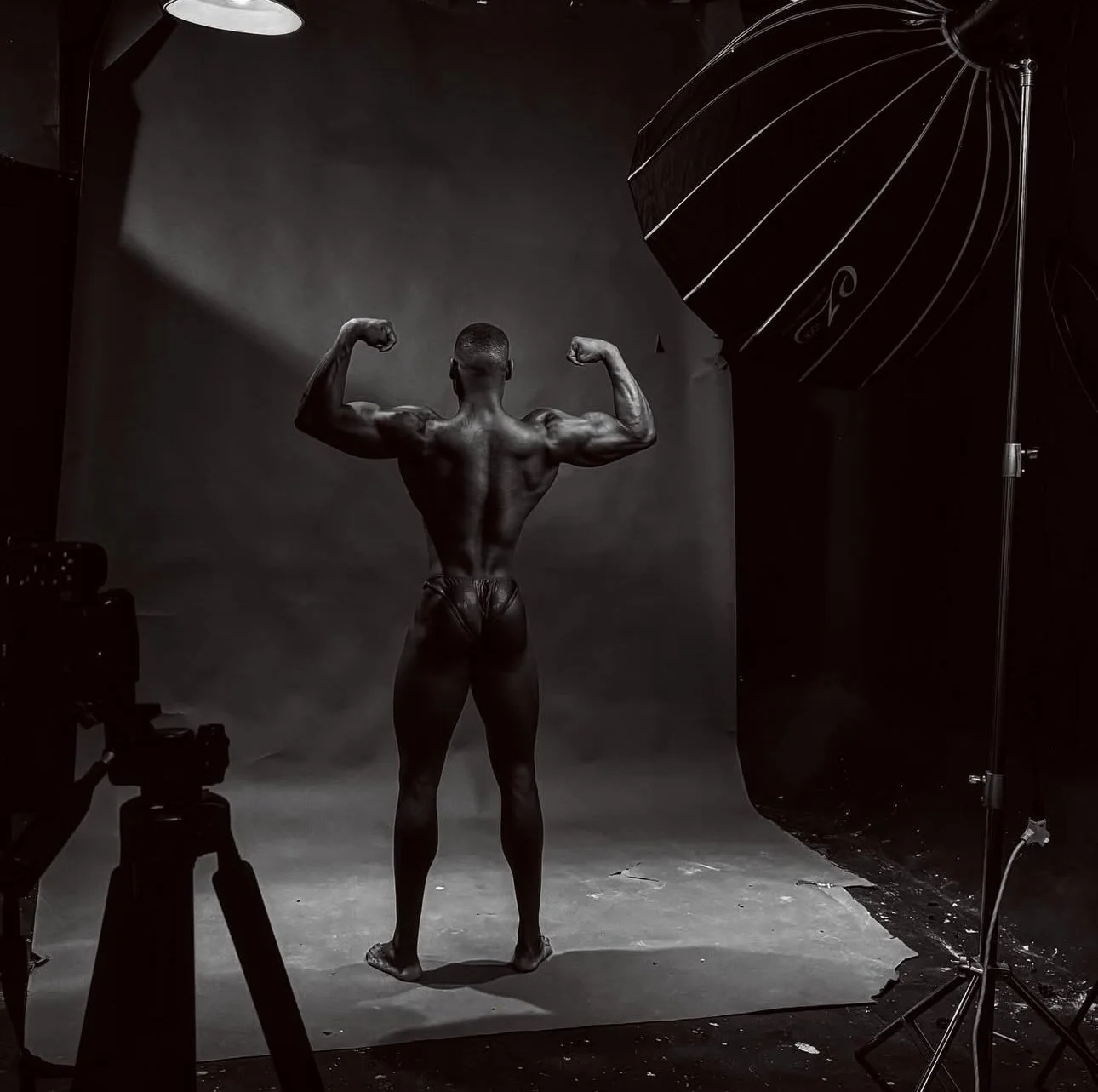 A male bodybuilder with muscular physique posing in a photography studio, back facing the camera, with studio lighting and equipment visible.