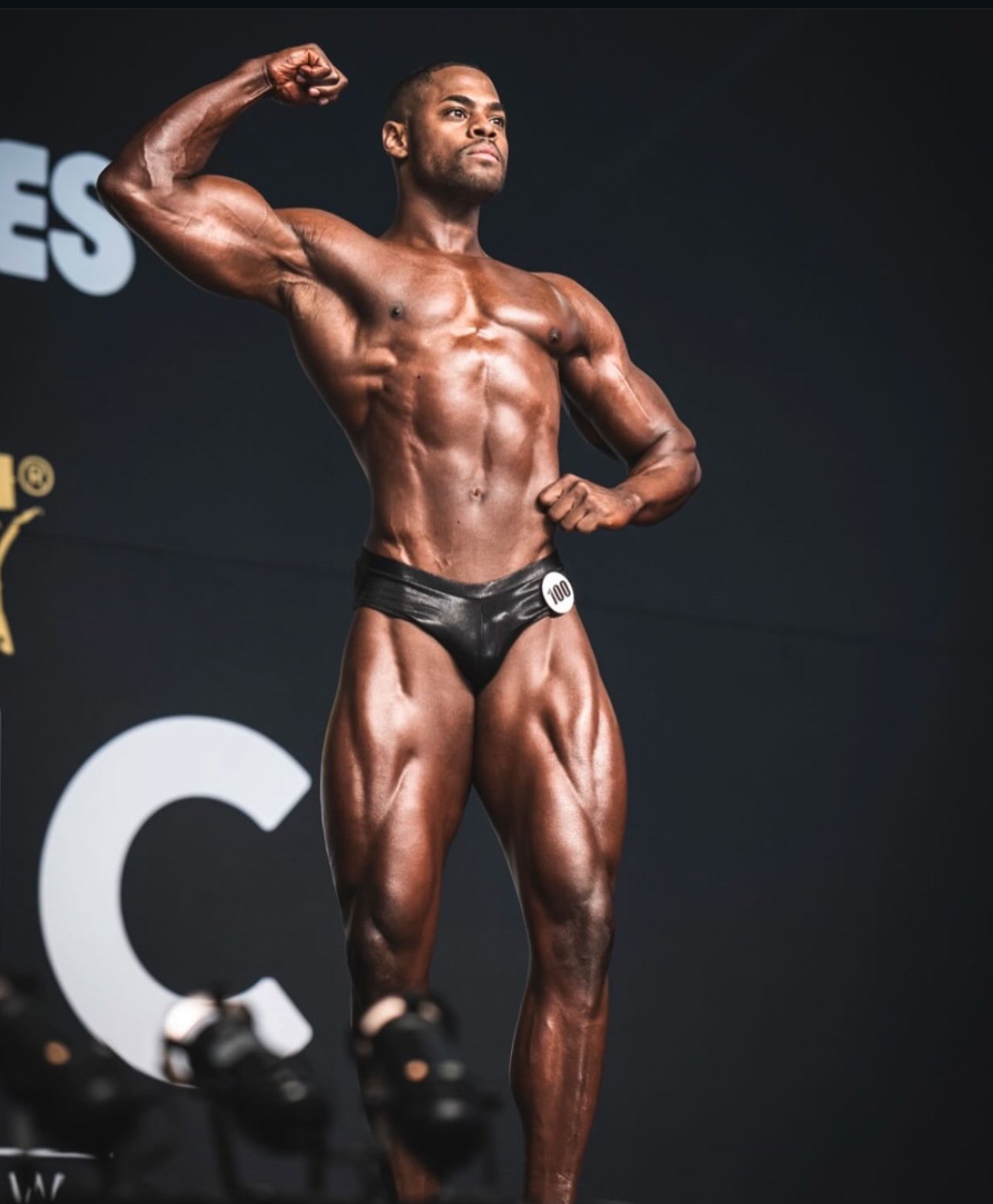 A muscular male bodybuilder with dark skin in a pose on stage, flexing his biceps and chest, wearing black posing trunks with the number 100.