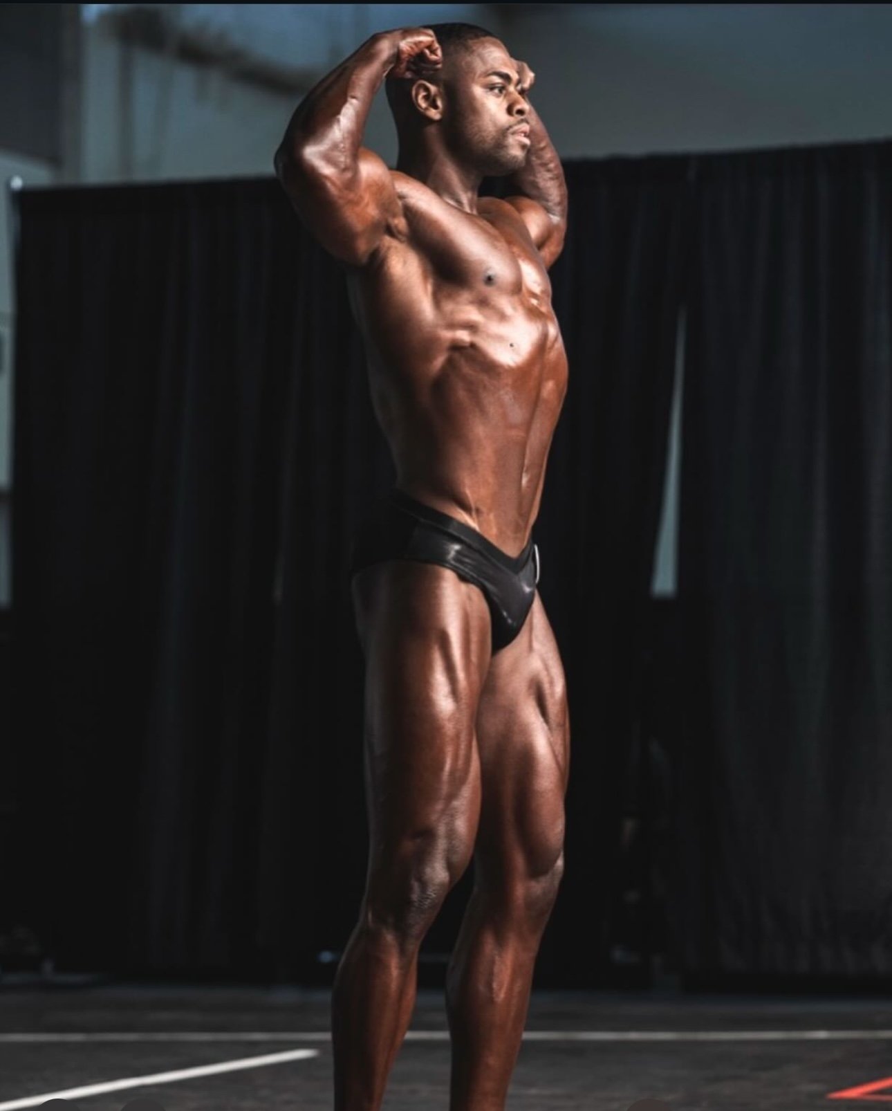 A male bodybuilder standing on stage with hands behind his head, showcasing his muscular physique.