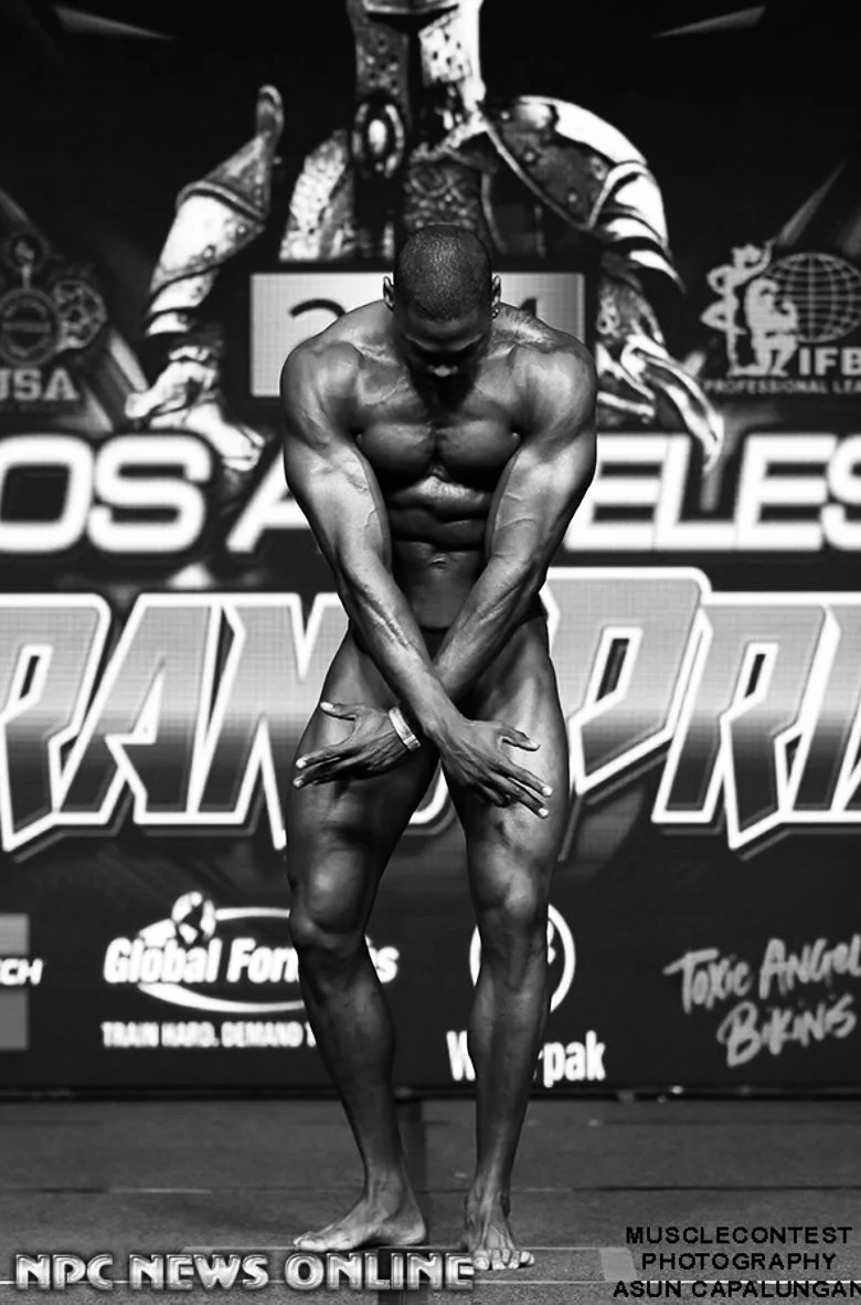 Black and white photo of a male bodybuilder on stage during a competition, posing with his arms crossed over his lower abdomen and looking down, with logos and text in the background.
