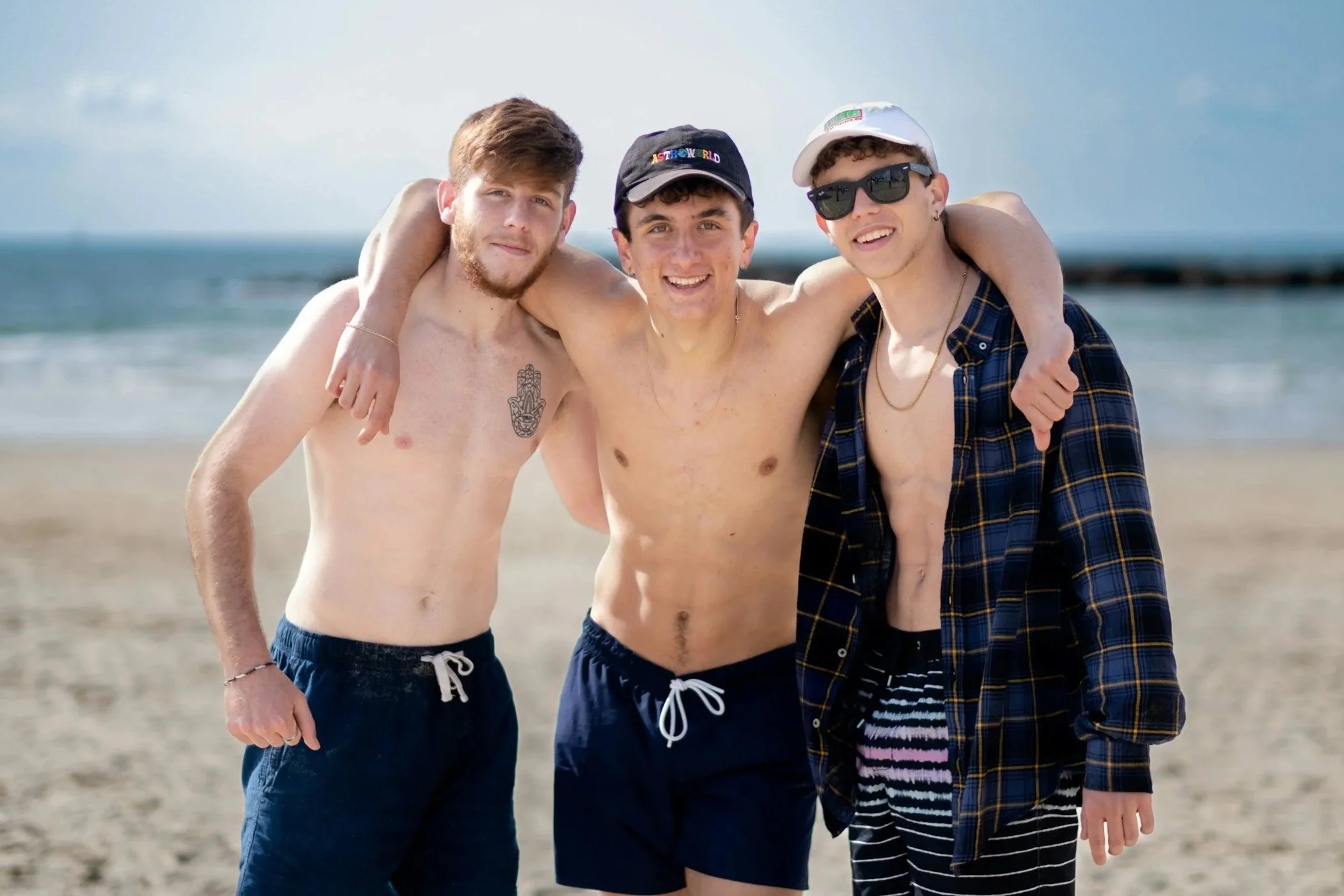 three white young men on the beach with blue sea behind them