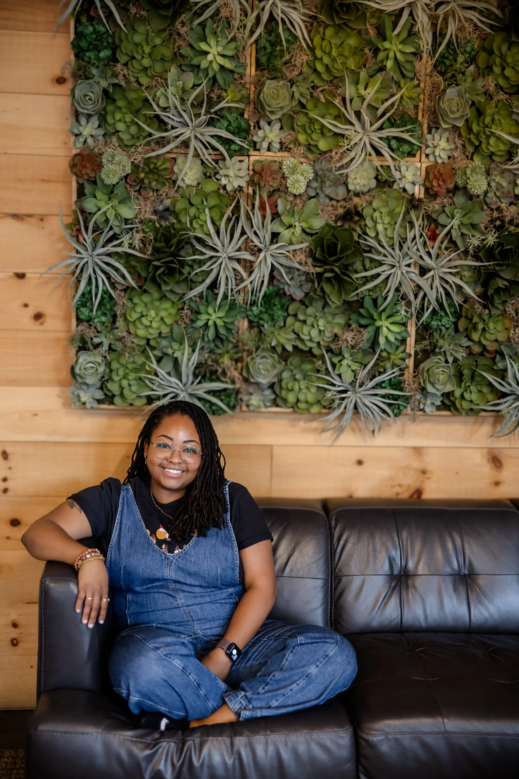 A black woman with glasses and locs sitting on a black leather couch in front of a wall decorated with a large green succulent plant arrangement. doula, birthworker, Massachusetts, Boston, herbalist, plants, advocate