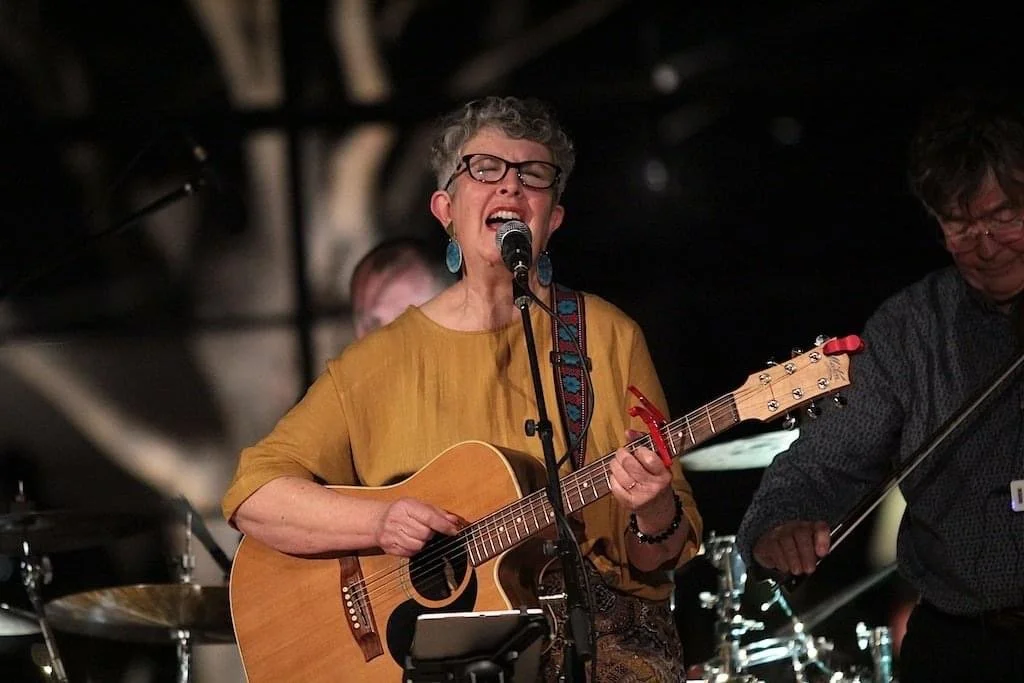 A woman with short curly hair, glasses, and turquoise earrings is singing and playing an acoustic guitar on stage. She is wearing a mustard-colored top and appears to be passionately performing.