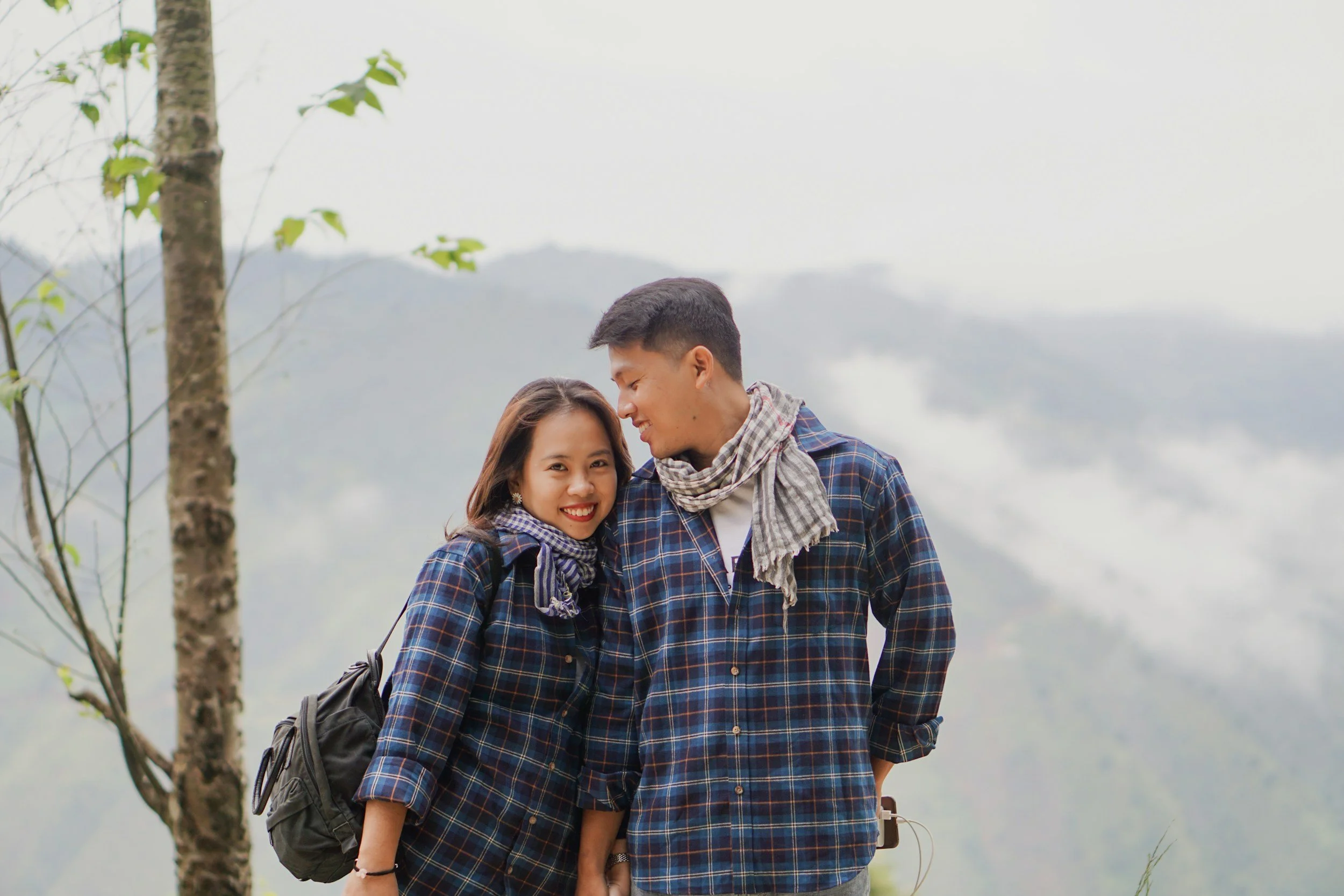 A smiling couple dressed in plaid shirts with scarves, standing outdoors in a mountainous, foggy landscape, sharing an affectionate moment.