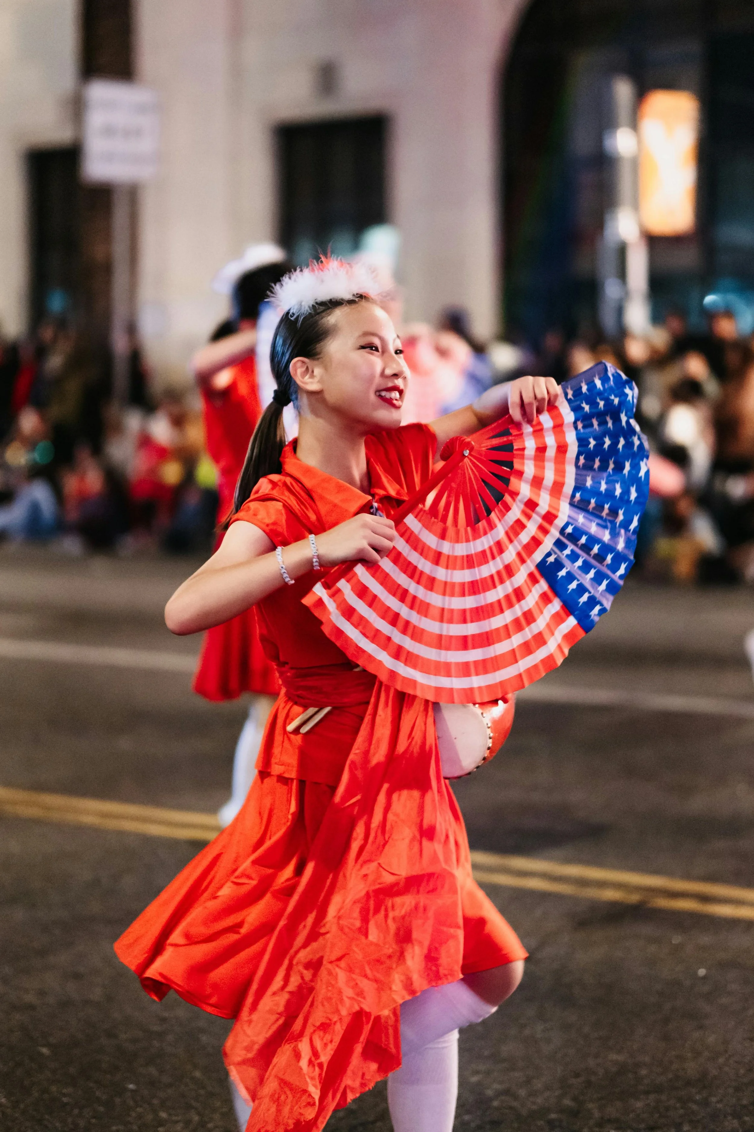A woman dressed in a red costume is holding an American flag-themed fan during a parade at night. She is smiling and wearing a fluffy white and pink headband, with a crowd of spectators in the background.