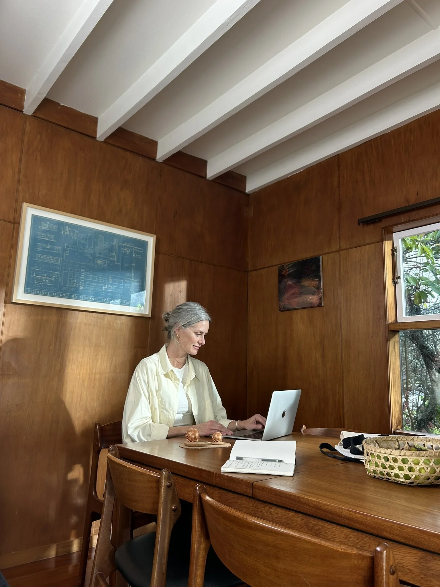 An elderly woman with gray hair working on a laptop at a wooden dining table in a room with wood-paneled walls and a window showing greenery outside.