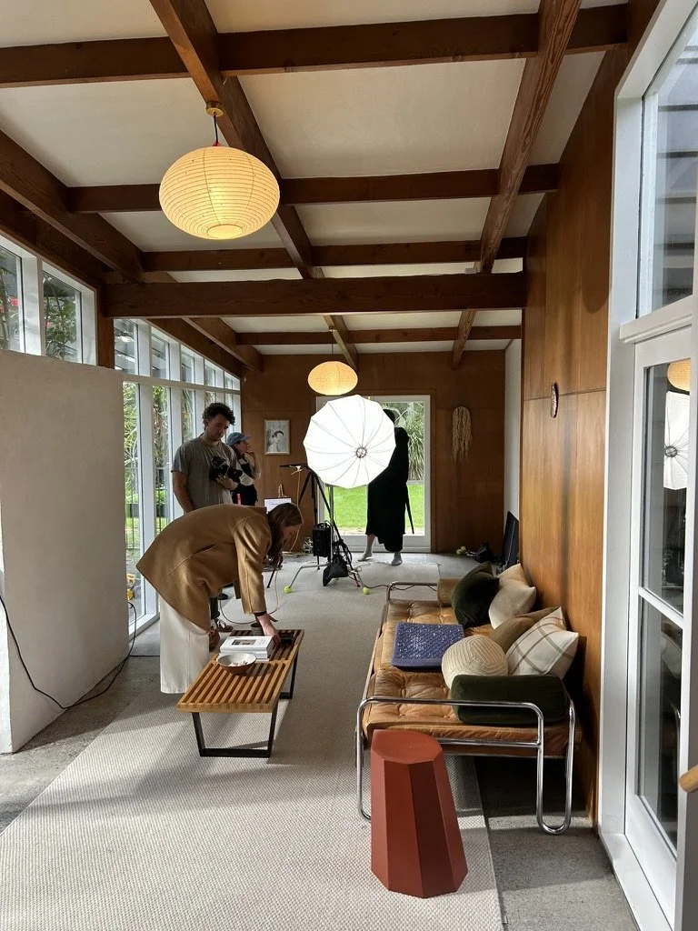 Interior of a wooden room set up for a photo shoot with large windows, a brown leather couch with pillows, a small table with equipment, and professional lighting with umbrellas, with people preparing for the shoot.