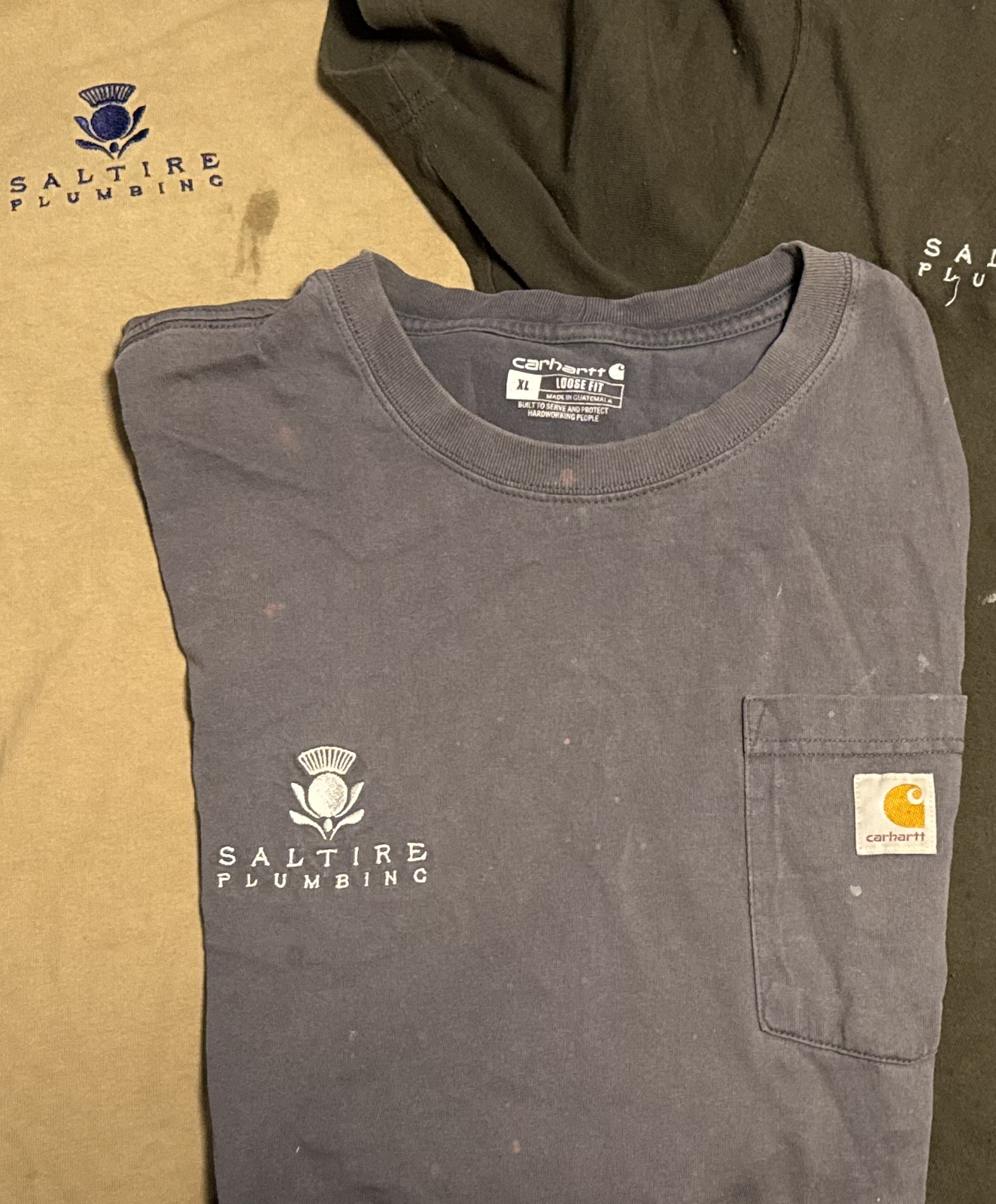 A folded dark gray Carhartt t-shirt with a small pocket on the front, displaying the Saltire Plumbing logo in white and purple, placed on top of a brown Saltire Plumbing shirt and a dark green Saltire Plumbing shirt, all with the Saltire Plumbing log