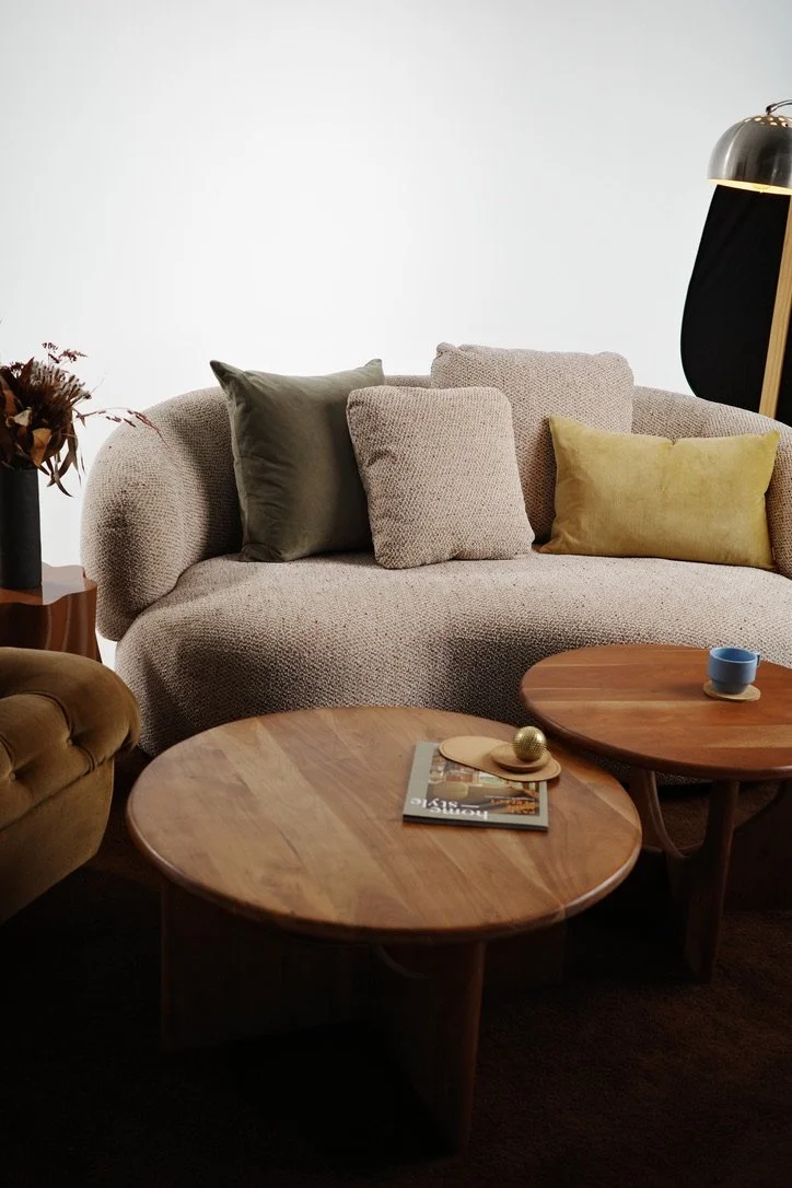 Living room with beige textured sofa, assorted cushions, a wooden coffee table with magazines and decor, a side table with a flower arrangement, and a black floor lamp with a gold interior shade.