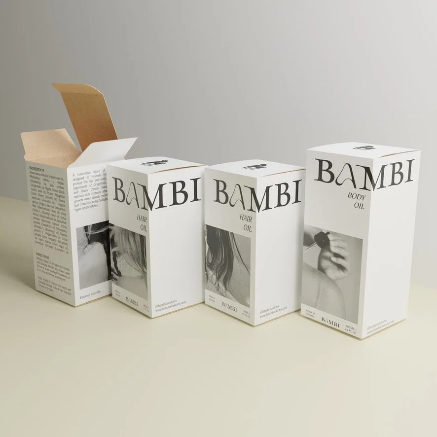 Four white boxes labeled Bambi hair and body oil, with one open box revealing interior packaging, aligned on a beige surface against a plain wall.