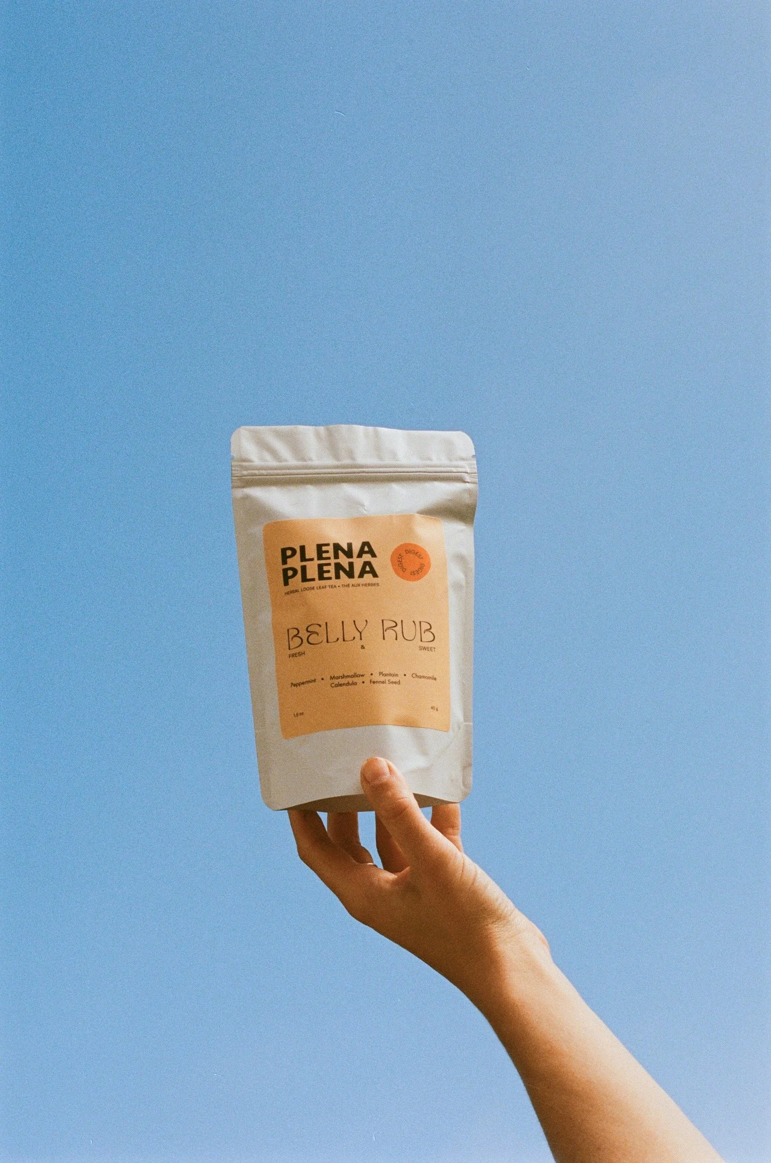 Hand holding a white package of Plena Plena belly rub herbal tea against a blue background.