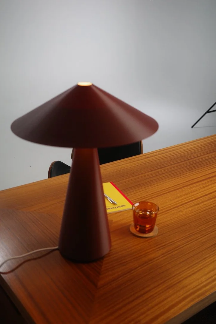 A wooden table with a brown mushroom-shaped lamp, a small amber glass, a yellow notepad with a pen, and a black chair in the background.