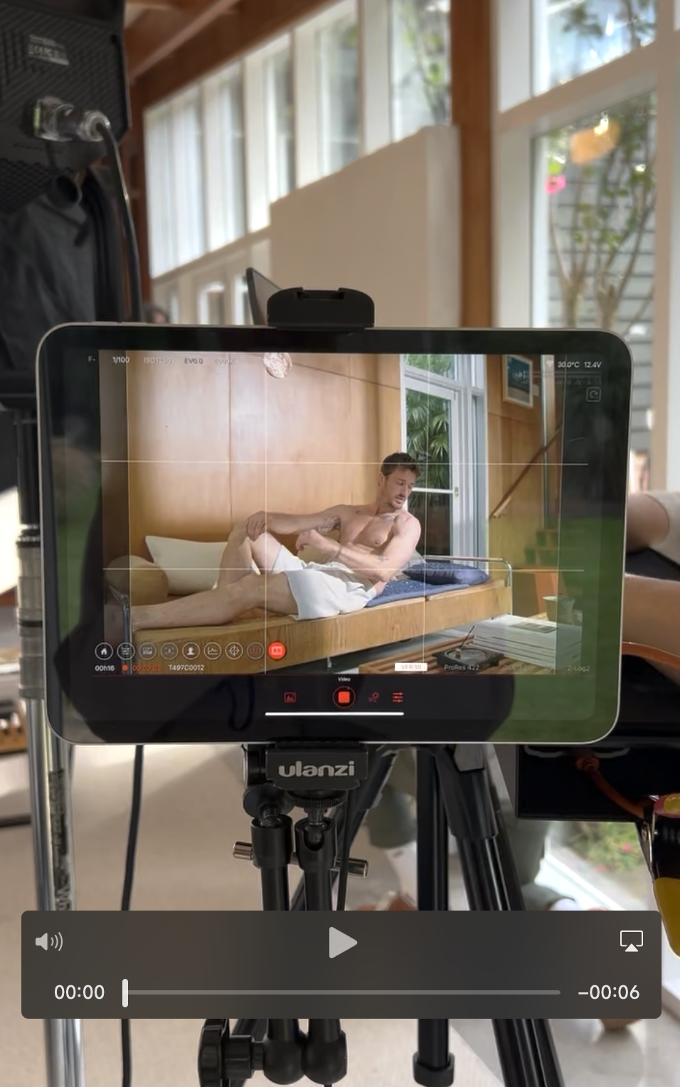 A camera on a tripod recording a shirtless man lying on a bed with a wooden headboard inside a bright room with large windows and plants outside.