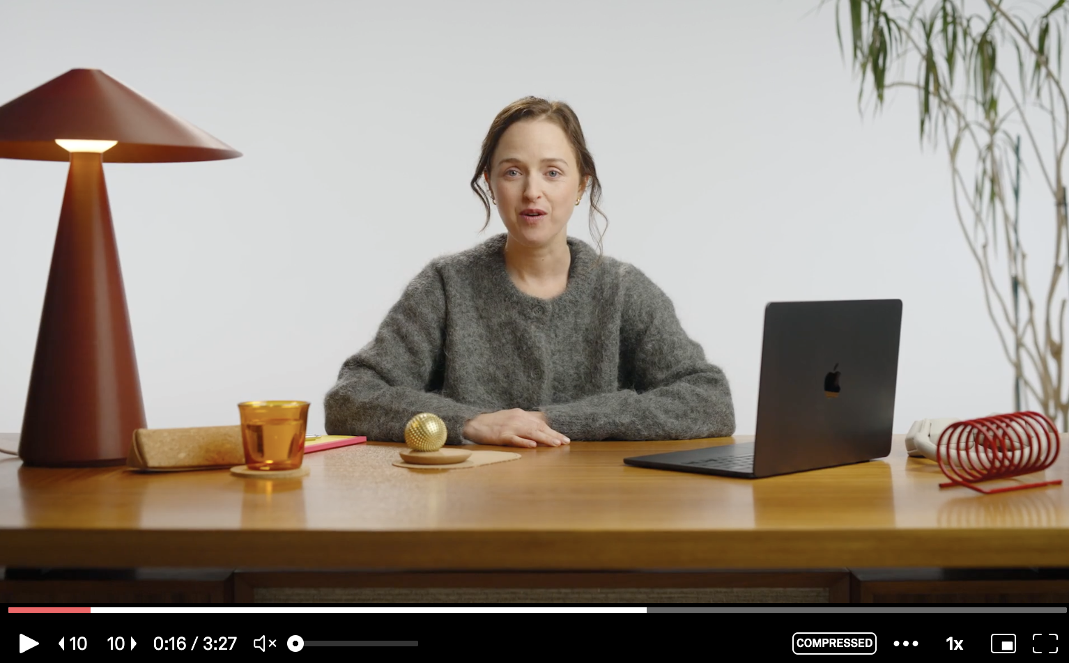 A woman with brown hair wearing a gray sweater sitting at a wooden desk with a black laptop, a yellow glass, a gold decorative object, a pink notebook, a tan brick, and a red desk organizer in a room with white walls and a potted plant behind her.