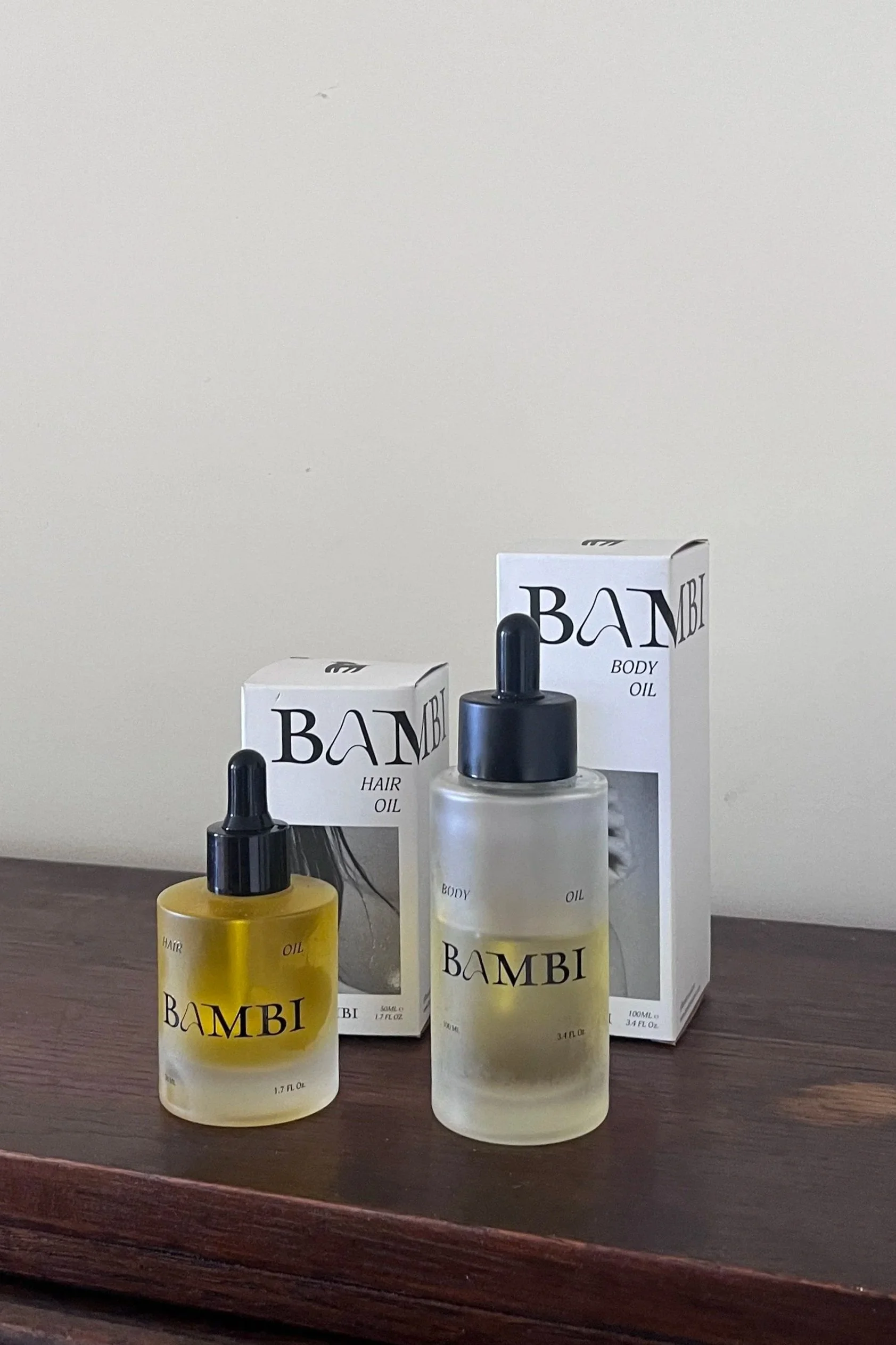 Two bottles of BAMBI body oils, one labeled 'Hair Oil' and the other 'Body Oil,' placed on a wooden surface with their respective packaging boxes in the background.