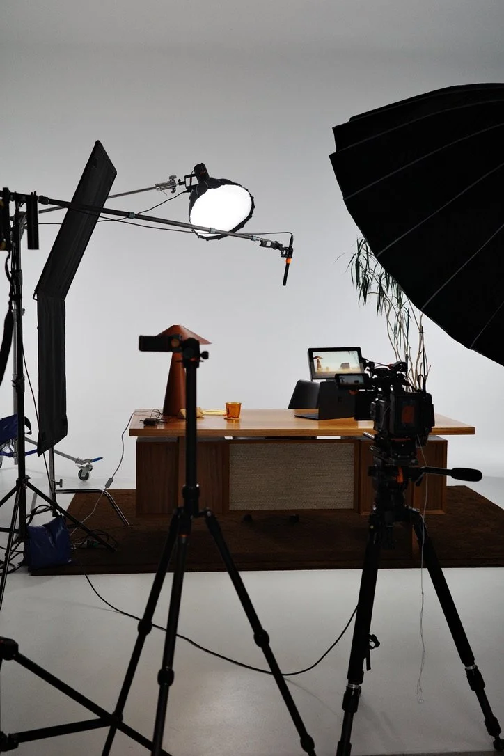 Photo of a filming or photography studio setup with a wooden desk, a computer, a small orange board, and a glass on the desk. Studio equipment including a large umbrella light, a softbox, and a reflector are visible, along with a potted plant in the