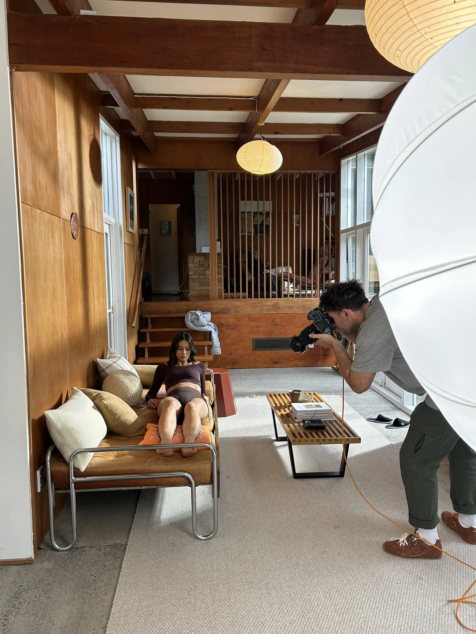 A woman lying on a vintage brown sofa with beige pillows in a room with wooden walls and ceiling beams, being photographed by a man with a camera near a large window, with another person sitting on a bench in the background.