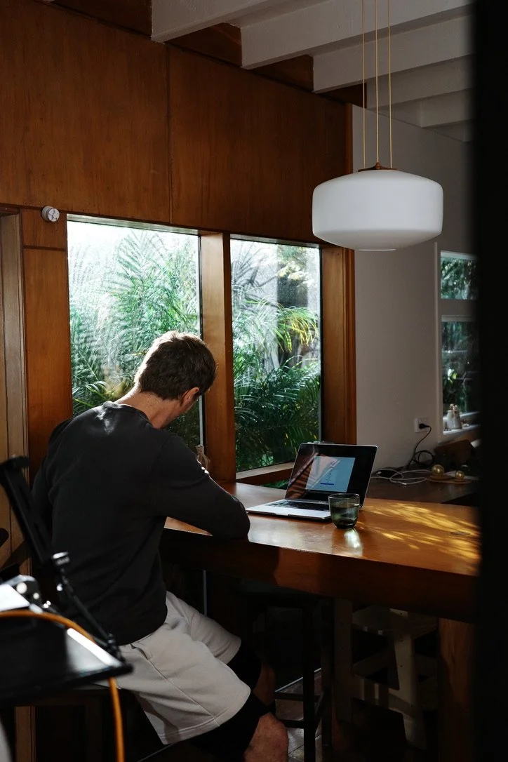 A person sitting at a wooden table using a laptop in a room with large windows showing greenery outside, with sunlight casting shadows inside.