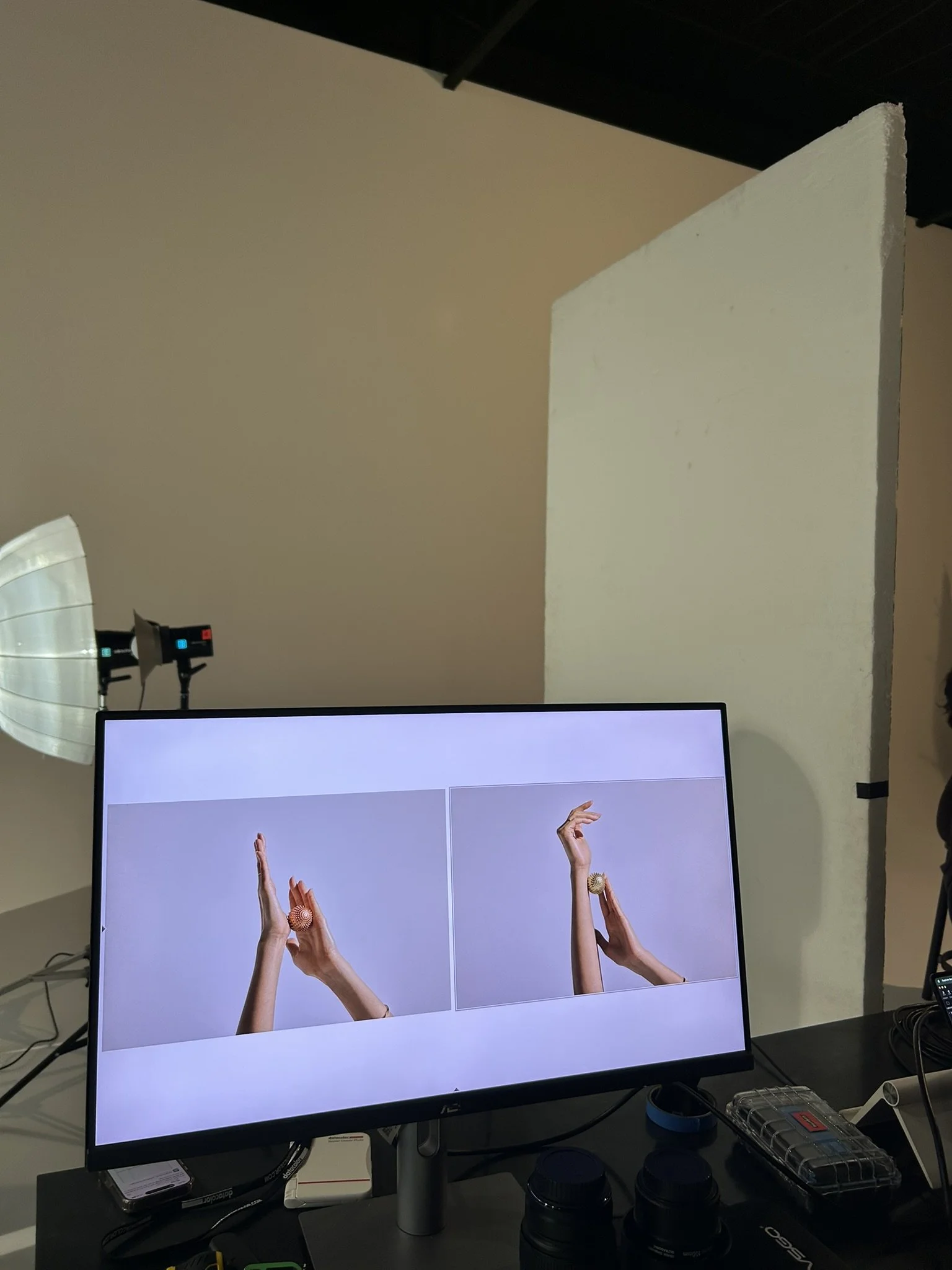 Setup for a photo shoot with a monitor displaying two images of hands holding textured objects. Studio lighting and a partition are visible in the background.