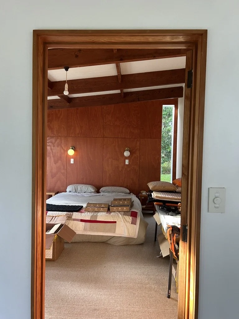 View of a bedroom through a doorway with wooden trim. The room has a bed with white bedding and two pillows, with books and items on top. There are two wall-mounted lights above the bed. To the right, there is a small window showing greenery outside,