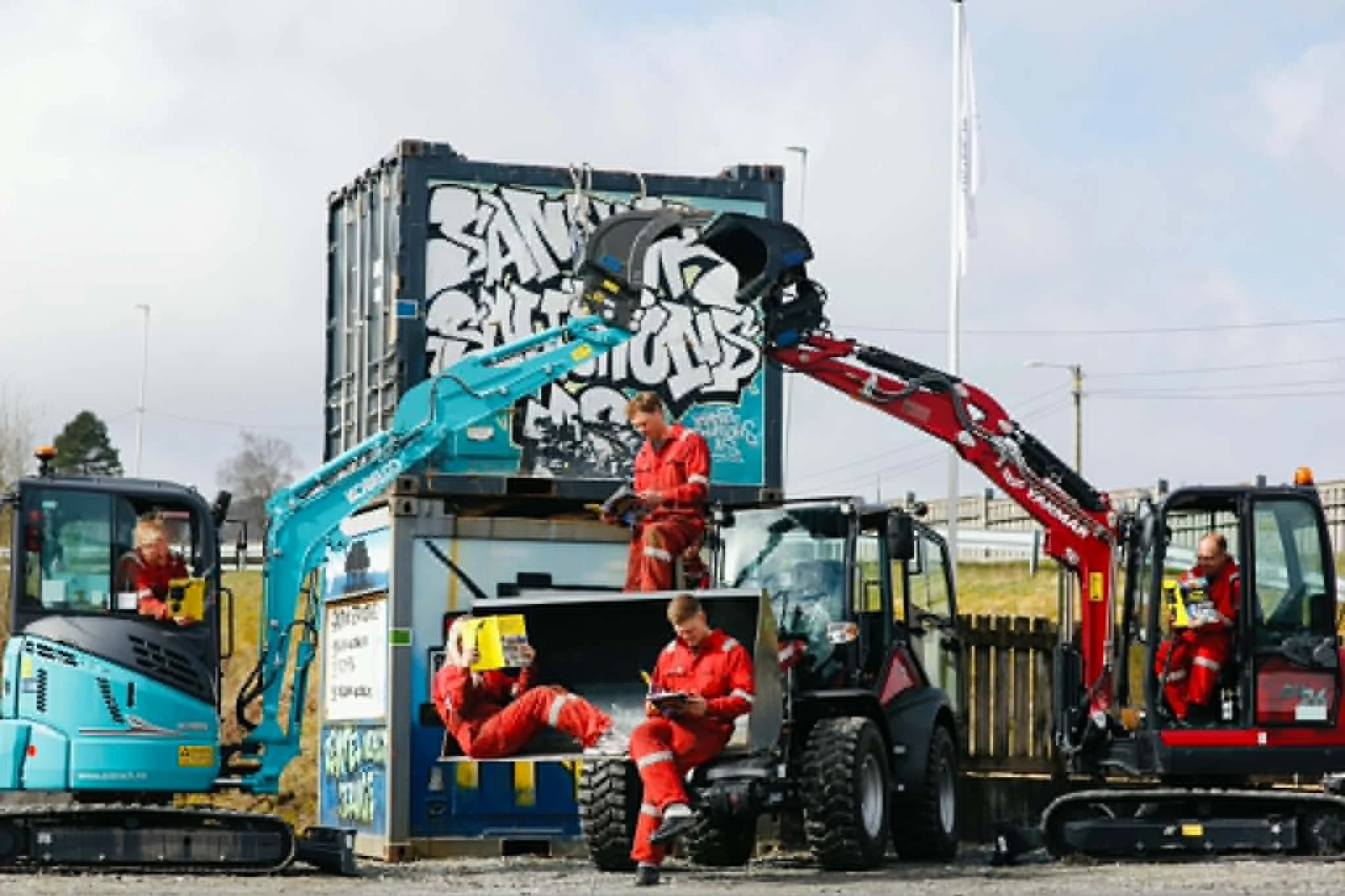 To gravemaskiner med operatører i arbeidsområde med personer i røde jobbekledninger, en av dem er sittende og leser, en annen står ved siden av. Bakgrunn med skilt og himmel.