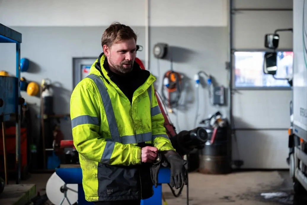 En mann i gul refleksvest med arbeidsutstyr i en verksted eller lager.